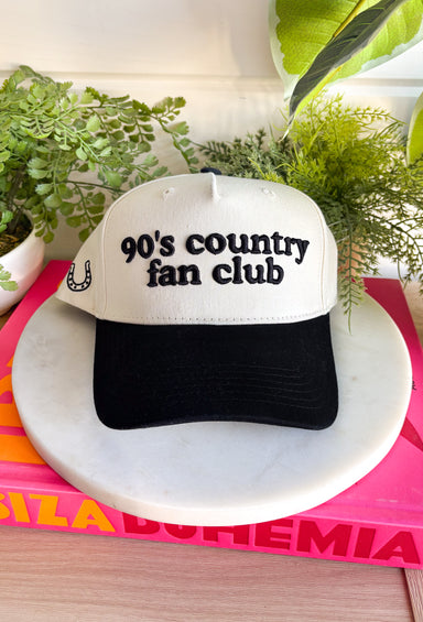 Cue the boot scootin’ playlist! This neutral trucker hat with a bold black bill brings all the throwback country vibes. Featuring “90’s Country Fan Club” across the front and a lucky horseshoe detail on the side, it’s the perfect nod to your favorite era of country music. With its classic trucker silhouette and easy neutral colorway, this hat pairs effortlessly with graphic tees, denim, and cutoffs for that laid-back, off-duty look.