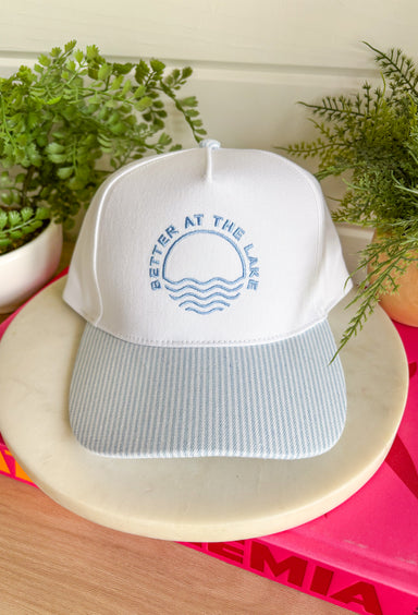 This trucker hat was made for sunny lake days and summer weekends! Featuring "Better At The Lake" embroidered across the front in light blue with a cute water design, it’s the perfect accessory for all your warm-weather adventures. The white crown paired with the blue, white, and off white striped bill gives it a fresh, coastal-inspired feel that looks just as good on the boat as it does grabbing lunch by the water. It’s an easy grab-and-go favorite you'll wear all season long. 