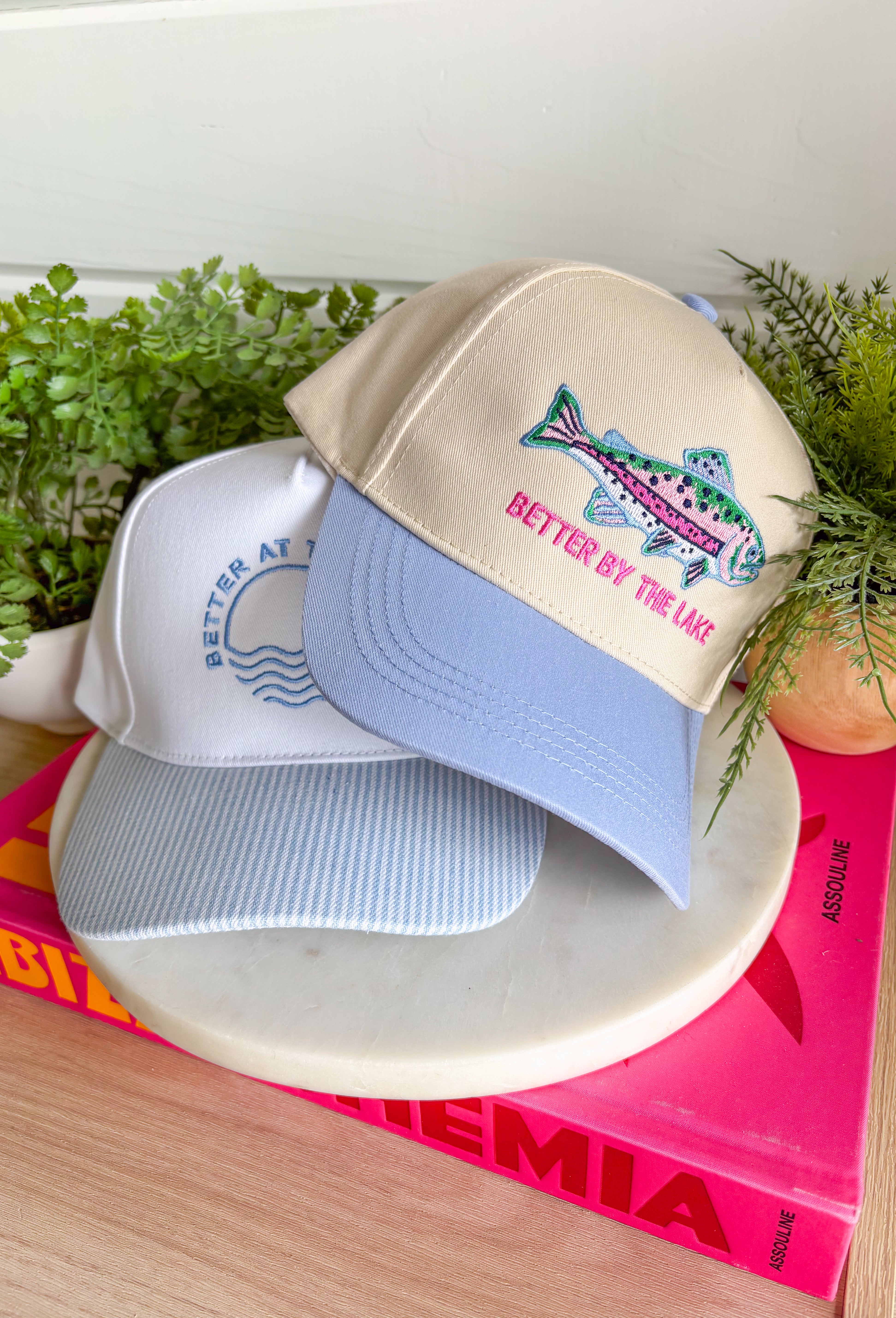 This trucker hat is made for long lake days, sunshine, and making memories by the water! Featuring a colorful embroidered fish and "Better By The Lake" stitched in bright pink, it brings all the fun summer vibes. The neutral crown keeps it easy to style, while the periwinkle bill adds the perfect pop of color. Whether you're spending the day on the boat, relaxing at the cabin, or heading out for a weekend getaway, this hat is the perfect finishing touch. 