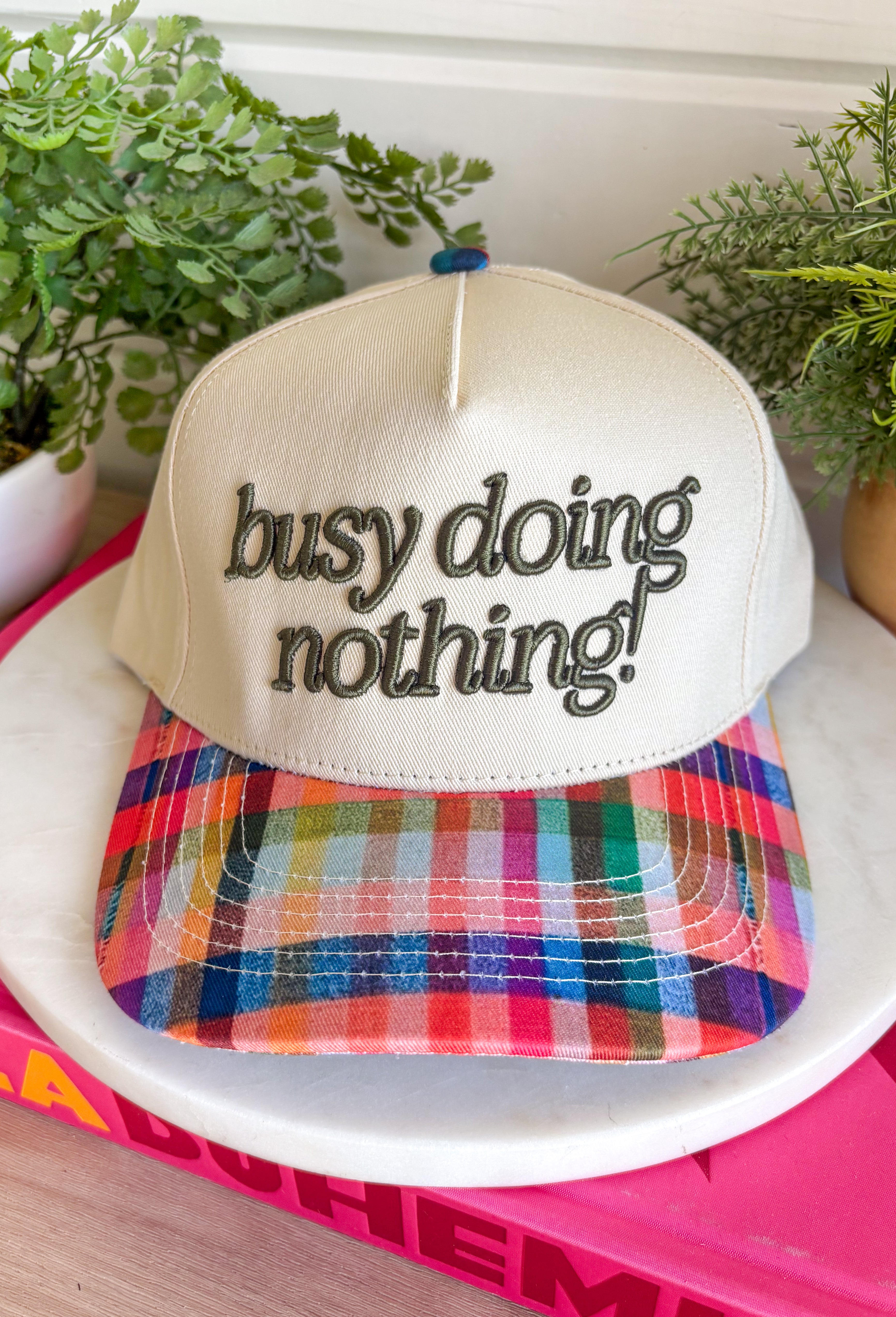 Some days are made for slowing down, and this trucker hat is here for it. Featuring "Busy Doing Nothing" embroidered in a rich deep olive color, it's the perfect accessory for cozy weekends, coffee runs, road trips, and laid-back fall adventures. The colorful plaid bill and matching top button add a fun seasonal touch, giving this hat plenty of personality while keeping it easy to style with your favorite casual outfits. 