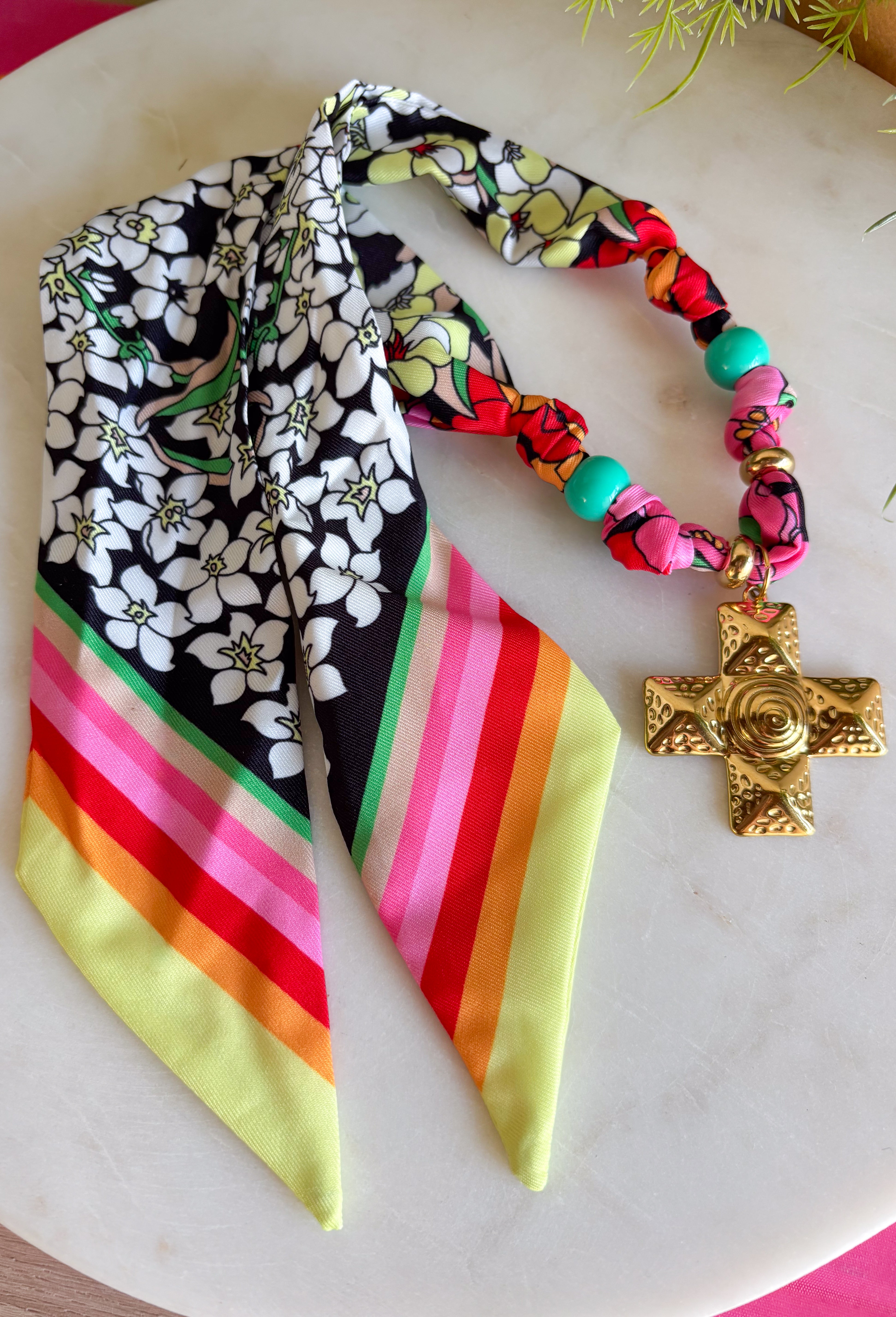 A little bold, a little western, and full of personality! This colorful bandana scarf necklace blends vibrant prints with meaningful details for a statement accessory you’ll love wearing. The scarf features black and white florals paired with bright stripes in pink, green, red, orange, and yellow for a fun pop of color. Finished with a gold textured cross pendant and a turquoise bead accent, this piece brings together playful style with a hint of western charm. 