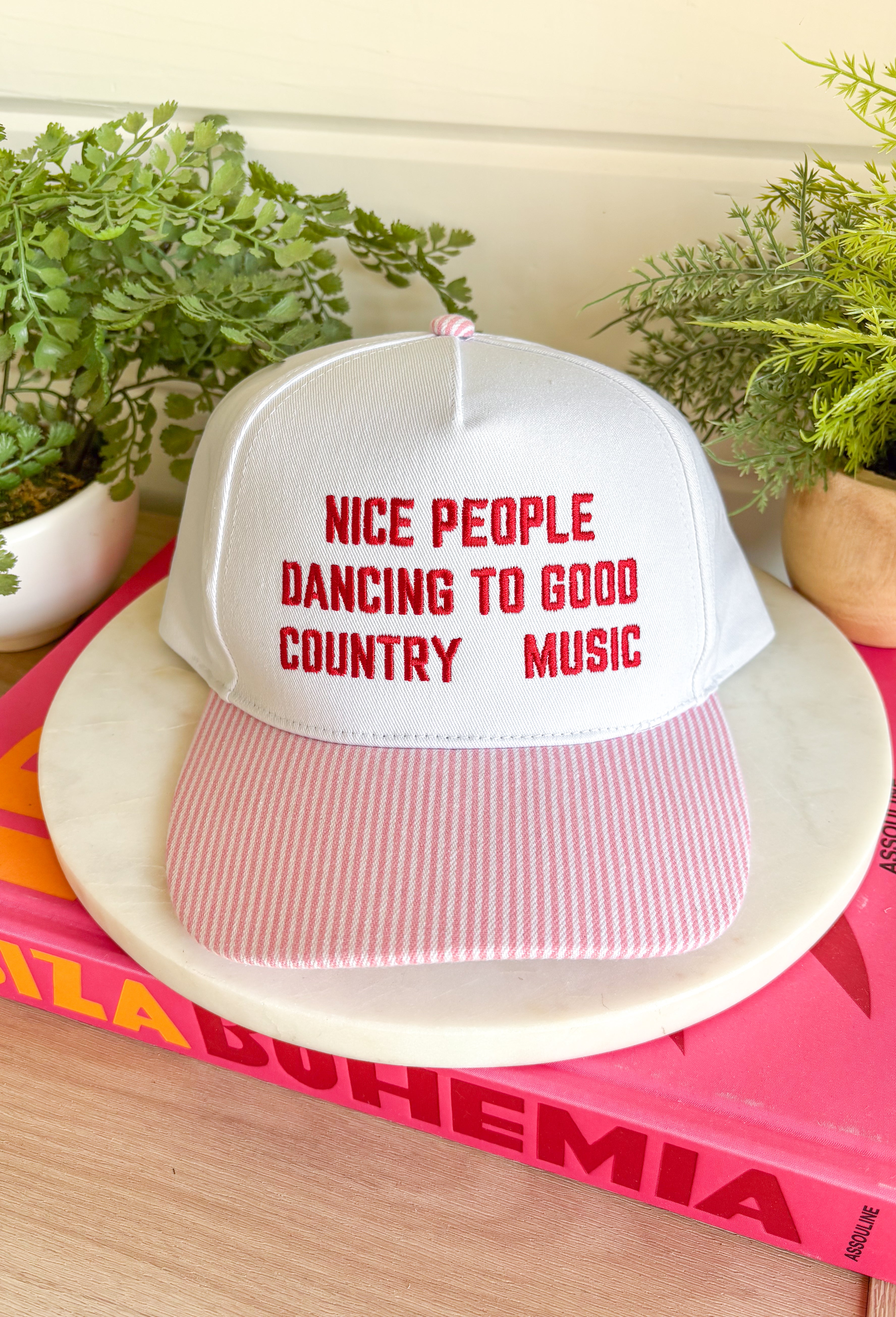 Cue the country playlist—this trucker hat was made for good times and great company! Featuring&nbsp;"Nice People Dancing To Good Country Music" embroidered across the front in bold red stitching, this playful hat is perfect for concerts, tailgates, rodeos, and weekends spent outdoors. The pink and white striped bill adds a fun pop of color, while the matching top button ties the look together. Finished with an adjustable fit, it's the perfect accessory for every country music lover.