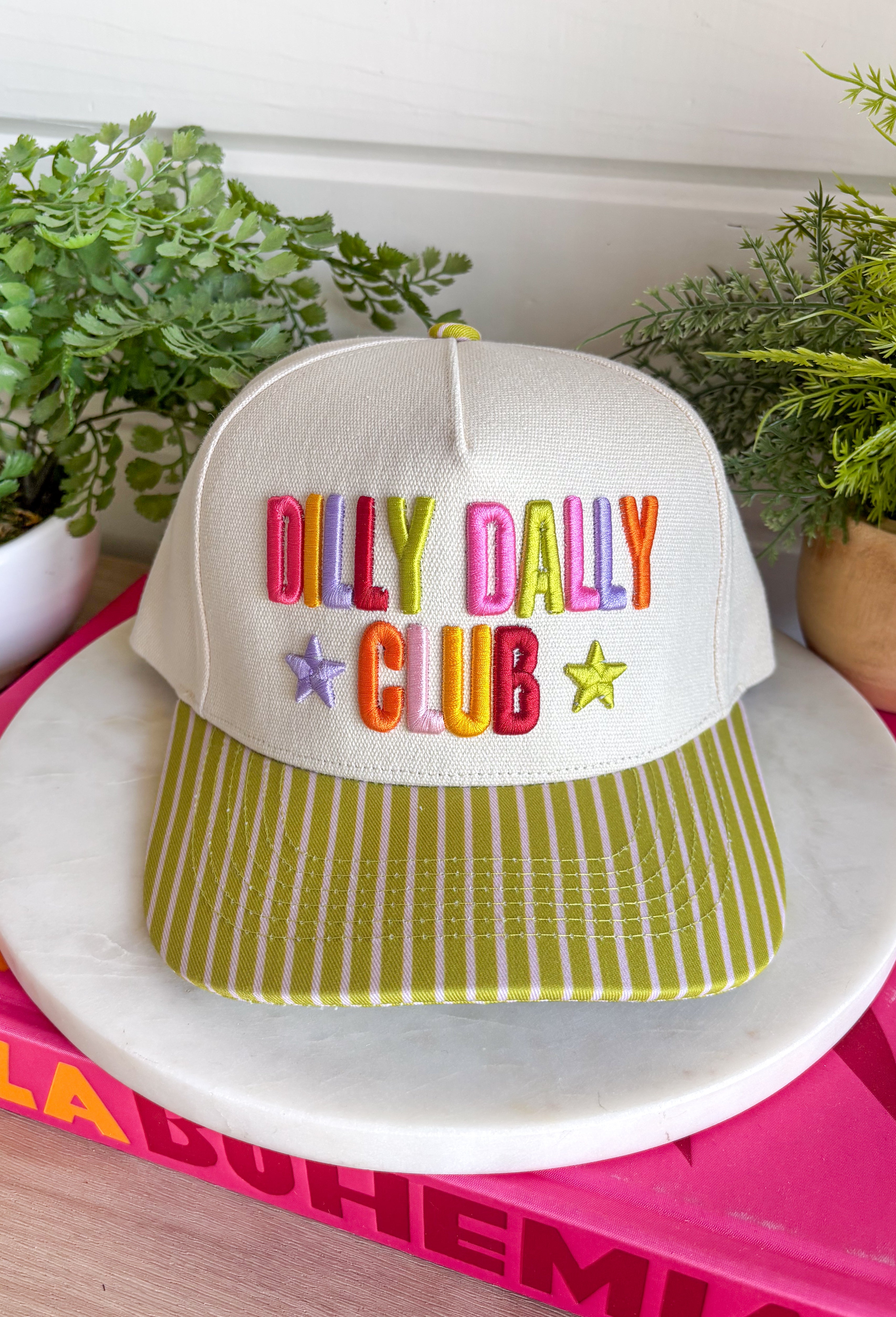 Slow down and enjoy the little moments with this colorful trucker hat. Featuring "Dilly Dally Club" embroidered in vibrant colors alongside a playful purple and lime green star, this hat is full of personality and happy vibes. The lime green bill with neutral stripe detailing and coordinating top button make it a standout accessory that's perfect for everything from weekend adventures to coffee runs and casual days out.
