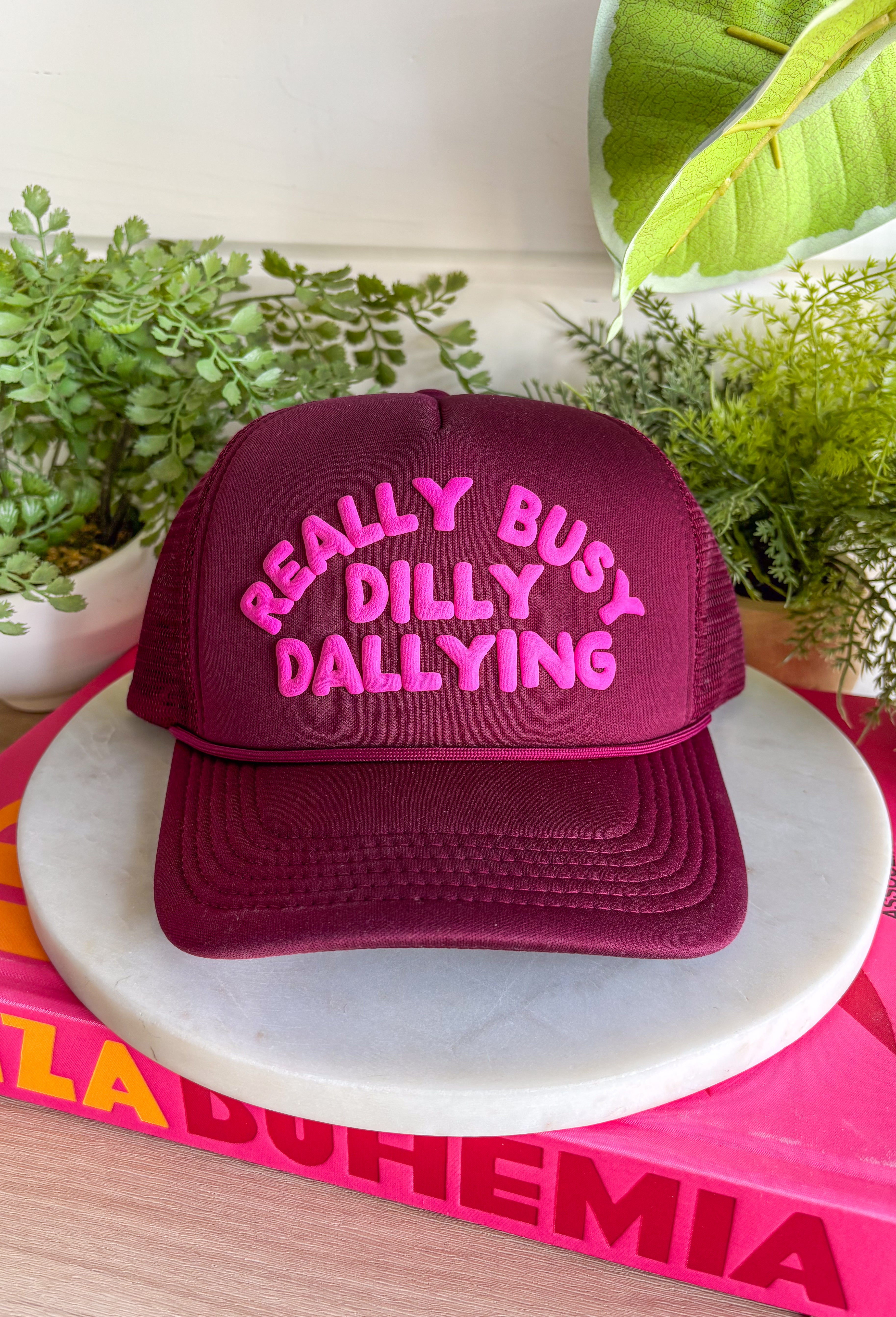This burgundy trucker hat features “Really Busy Dilly Dallying” in the cutest hot pink bubble font, giving it that fun, carefree personality we love. The bold color combo makes a statement while the classic mesh backing keeps it breathable and easy to wear. Toss it on for errands, ball games, coffee runs, or those days when you’re technically productive… just at your own pace.
