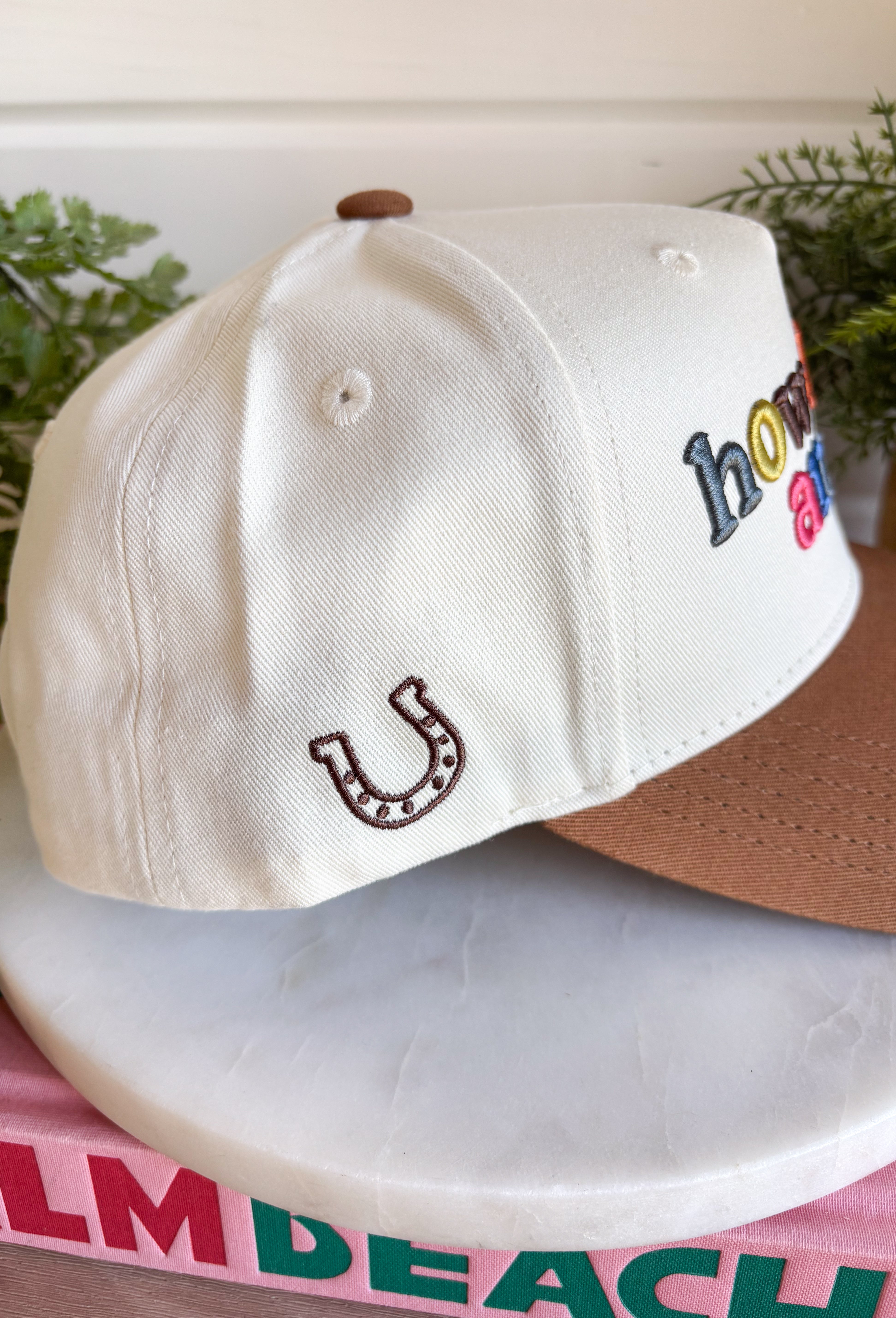 A little reminder to appreciate the good stuff! This neutral trucker hat from Etta &amp; East features colorful embroidered “How Lucky Are We” lettering across the front for a playful pop against the classic neutral design. A rich brown bill gives it an autumn-ready feel, while an embroidered horseshoe on the side adds an extra lucky touch. Finished with an adjustable fit, it’s an easy grab-and-go accessory for football weekends, coffee runs, road trips, and everyday errands.