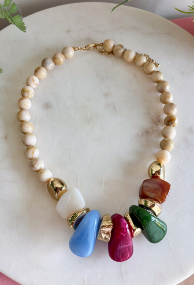 Neutral meets statement-making color with this eye-catching beaded necklace. A neutral beaded strand is accented with larger stone details across the front in rich brown, olive, fuchsia, and light blue, creating a playful mix of earthy and colorful tones. Gold beading between the stones adds a touch of shine and helps each color stand out. It’s the perfect piece when you want an accessory that’s easy to wear but still brings something extra to your outfit.