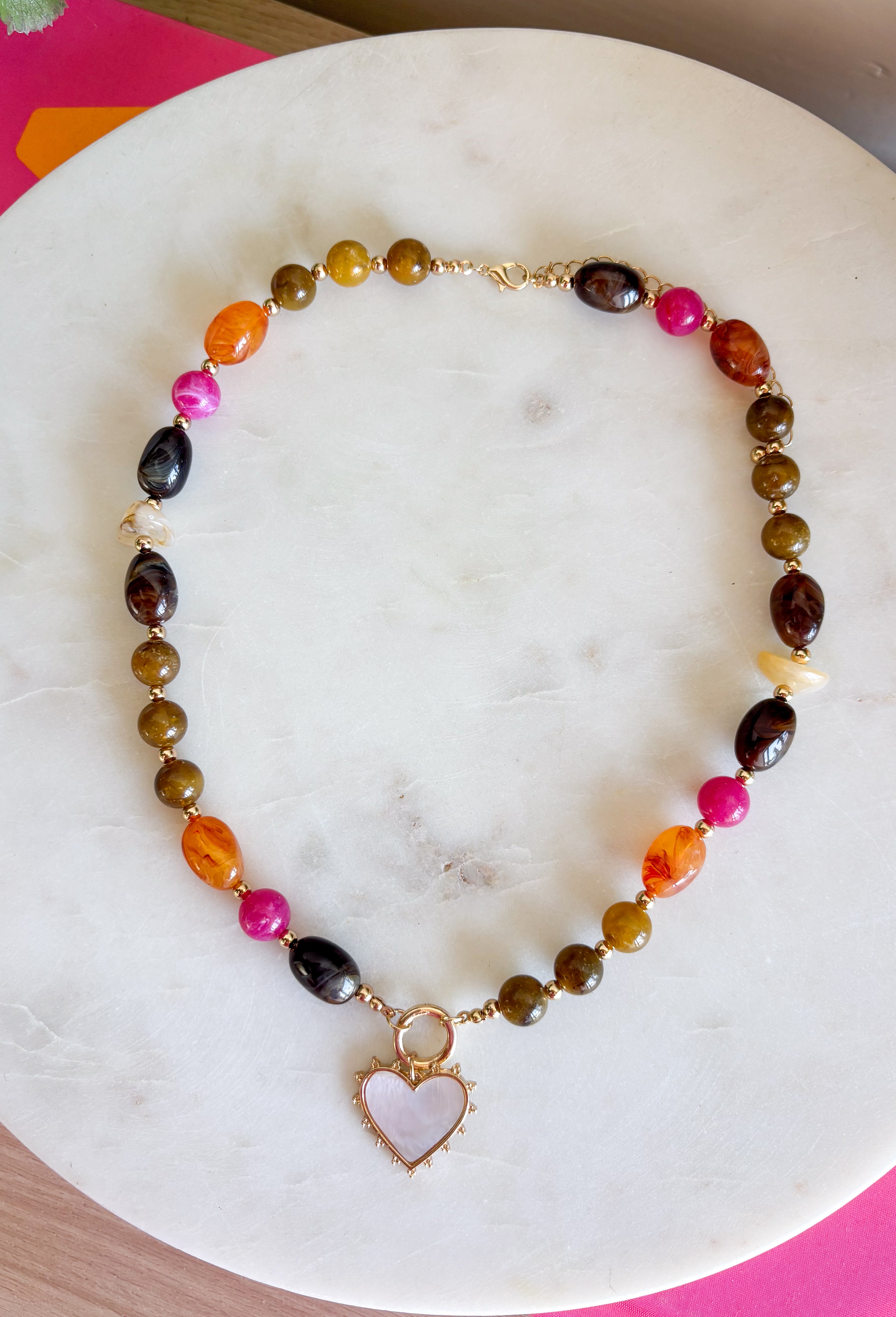 Rich autumn tones and elegant details come together in this stunning statement necklace. Featuring shades of brown with beautiful pops of pink and rust, this beaded design is accented with delicate gold spacer beads that add the perfect touch of shine. The mother-of-pearl heart pendant framed in gold creates a timeless focal point, while the toggle detail and lobster clasp complete this effortlessly elevated accessory. It's the perfect finishing touch for all your favorite fall looks.