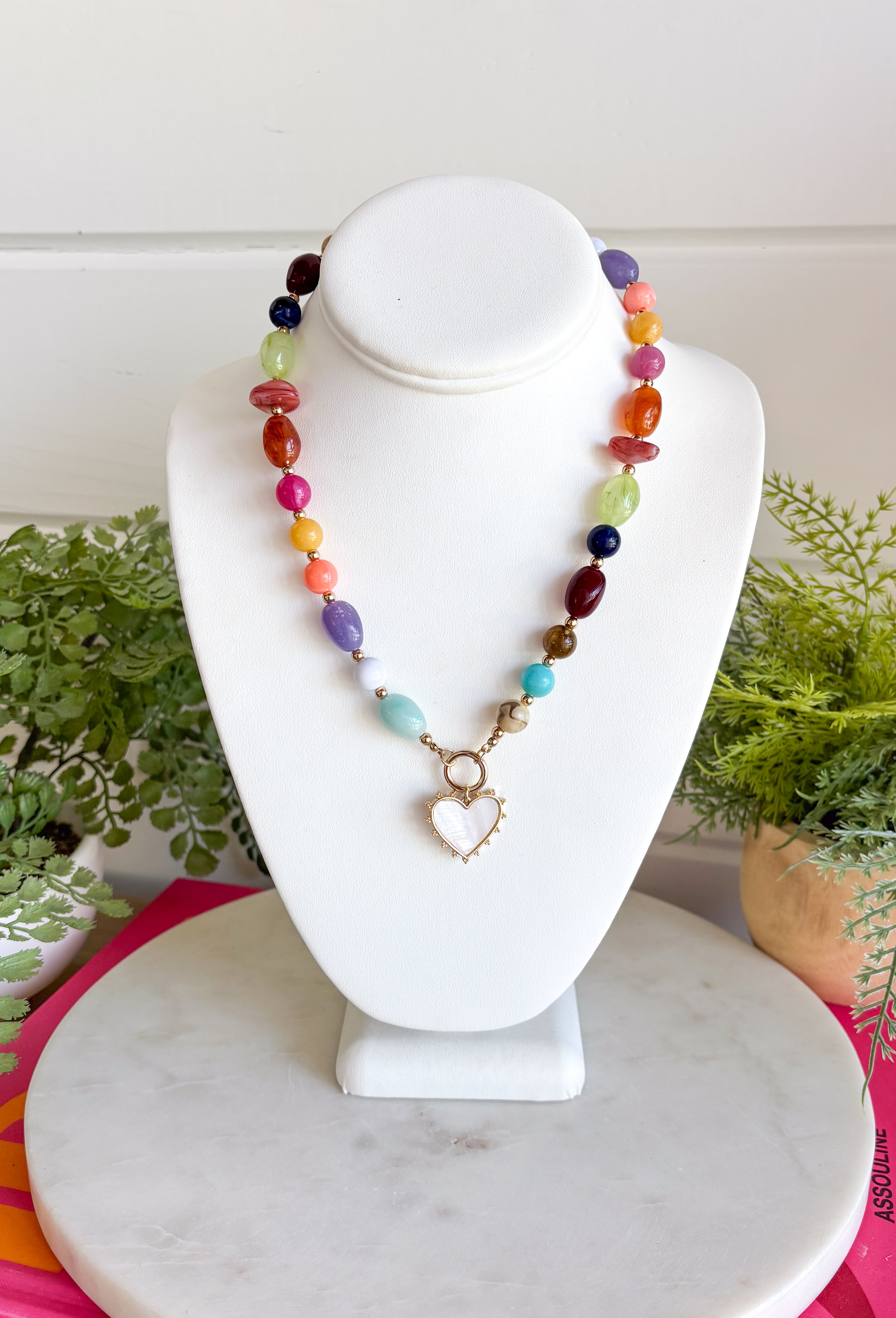 Rich autumn tones and elegant details come together in this stunning statement necklace. Featuring shades of brown with beautiful pops of pink, teal, green, purple, and rust, this beaded design is accented with delicate gold spacer beads that add the perfect touch of shine. The mother-of-pearl heart pendant framed in gold creates a timeless focal point, while the toggle detail and lobster clasp complete this effortlessly elevated accessory. It's the perfect finishing touch for all your favorite fall looks.