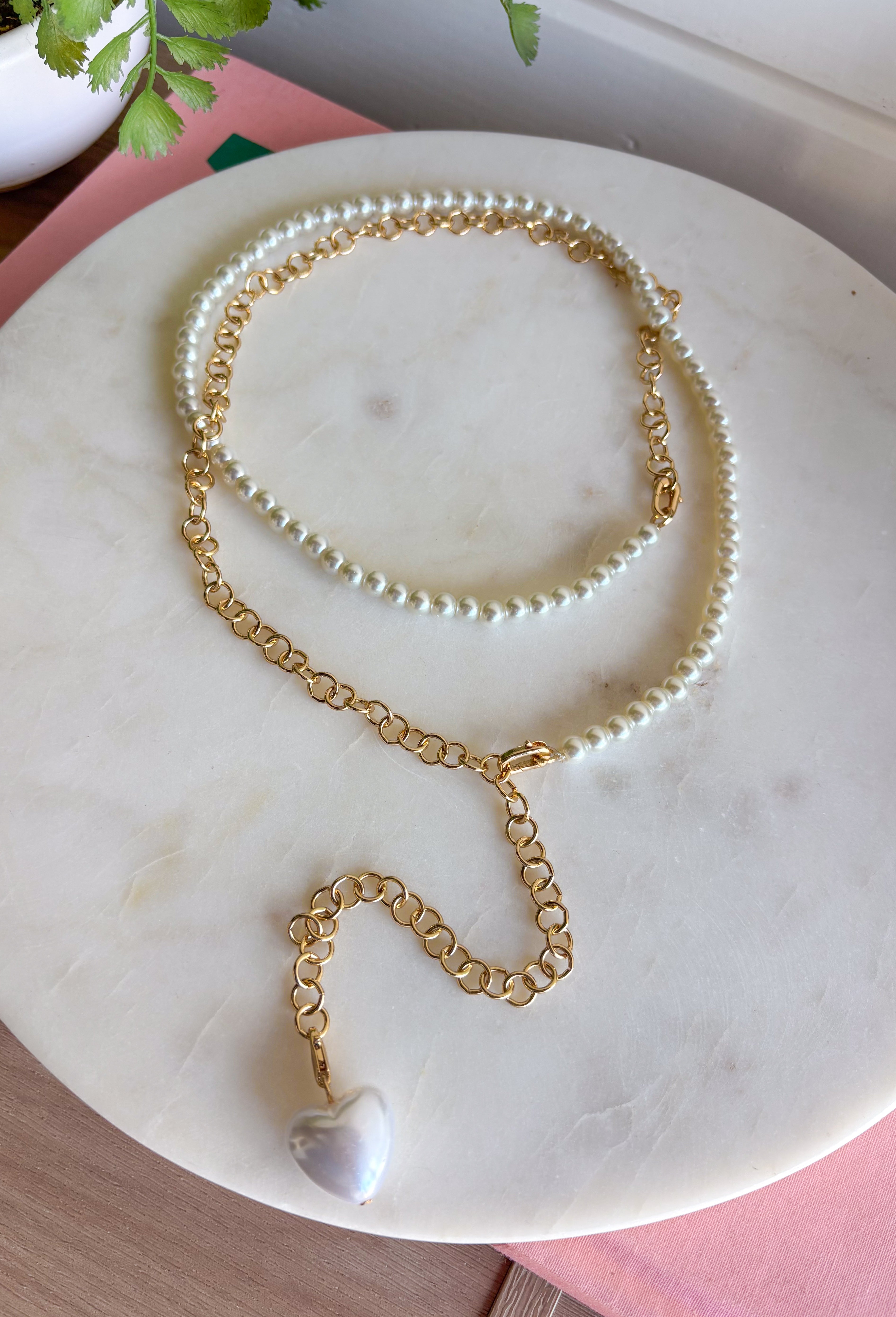 A layered necklace displayed on a beige fabric jewelry bust, featuring a gold-tone chain with a white heart pendant and a second layer of white pearl beads