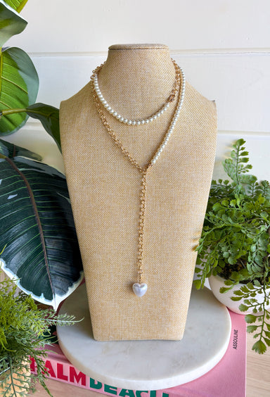 A layered necklace displayed on a beige fabric jewelry bust, featuring a gold-tone chain with a white heart pendant and a second layer of white pearl beads