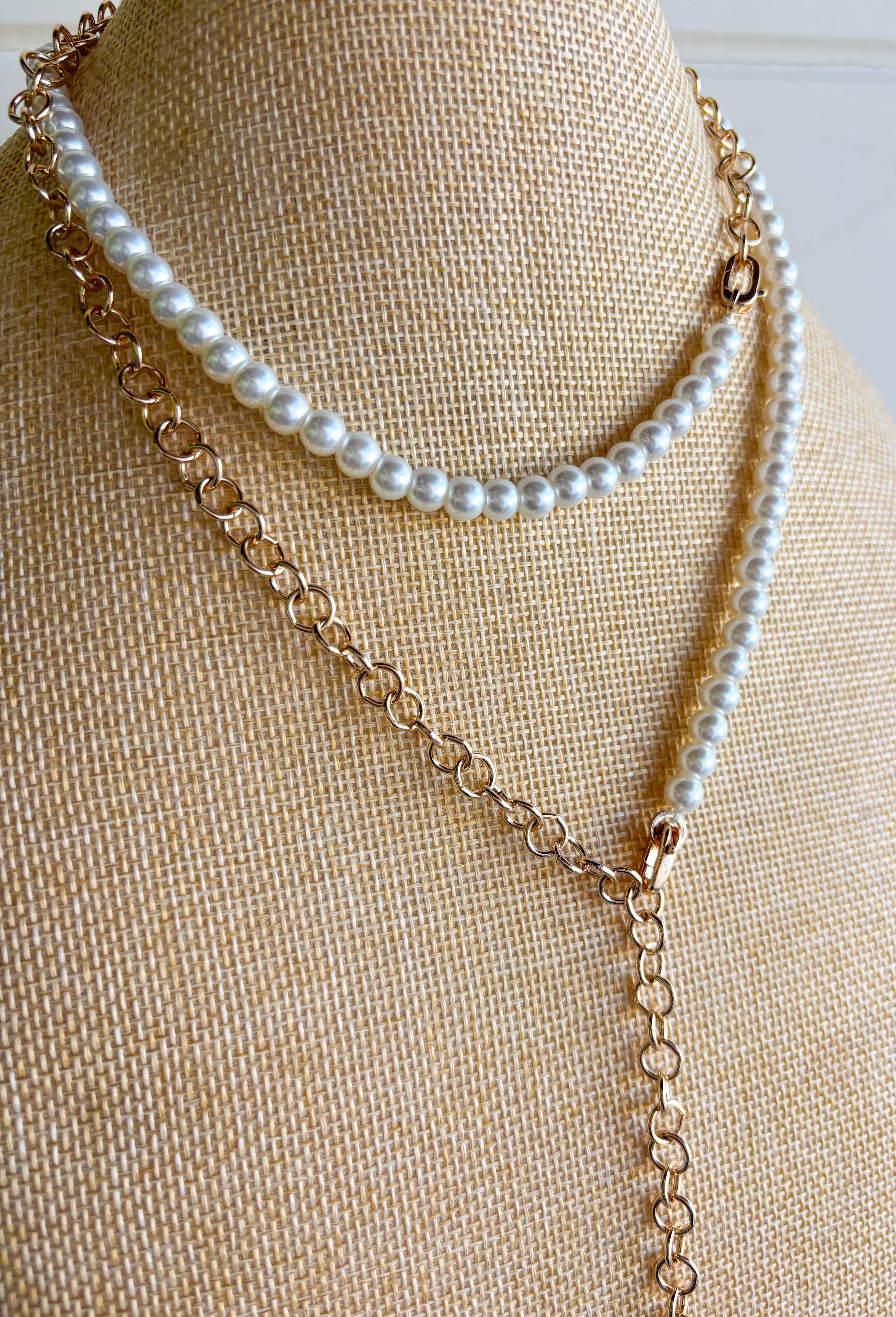 A layered necklace displayed on a beige fabric jewelry bust, featuring a gold-tone chain with a white heart pendant and a second layer of white pearl beads