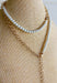 A layered necklace displayed on a beige fabric jewelry bust, featuring a gold-tone chain with a white heart pendant and a second layer of white pearl beads