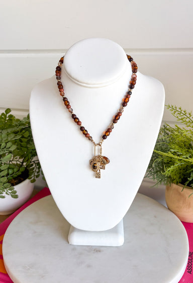 Rich earthy tones and meaningful details come together beautifully in this charm necklace that's perfect for the fall season. Featuring warm brown beads, this eye-catching piece is adorned with a mix of charms, including a gold cross, a natural brown stone, a sparkling brown crystal, and hammered gold accents for added texture and shine. Whether worn with a cozy sweater or your favorite dress, this unique necklace adds effortless charm and personality to every outfit.