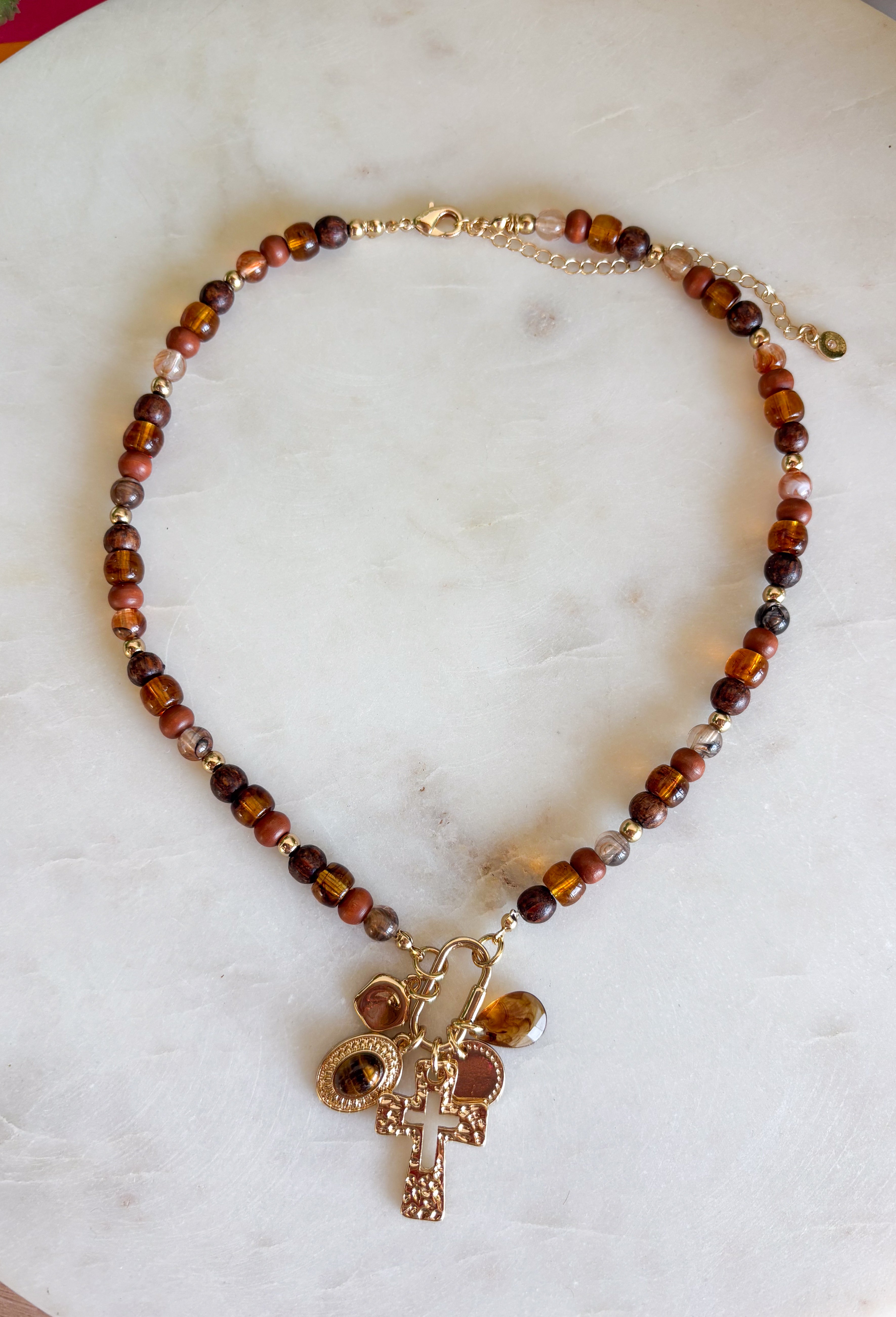 Rich earthy tones and meaningful details come together beautifully in this charm necklace that's perfect for the fall season. Featuring warm brown beads, this eye-catching piece is adorned with a mix of charms, including a gold cross, a natural brown stone, a sparkling brown crystal, and hammered gold accents for added texture and shine. Whether worn with a cozy sweater or your favorite dress, this unique necklace adds effortless charm and personality to every outfit.
