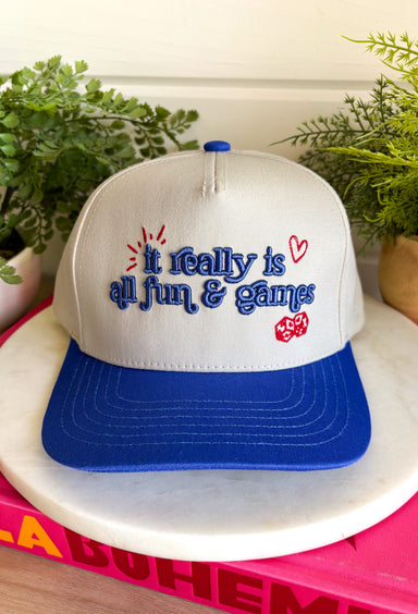 Life's too short not to have a little fun! This trucker hat features&nbsp;"It Really Is All Fun &amp; Games" embroidered in royal blue with red embroidered heart and dice details for the perfect playful touch. The neutral crown paired with the royal blue bill creates a classic color combination that's easy to style, while the fun graphic adds personality to every outfit. 