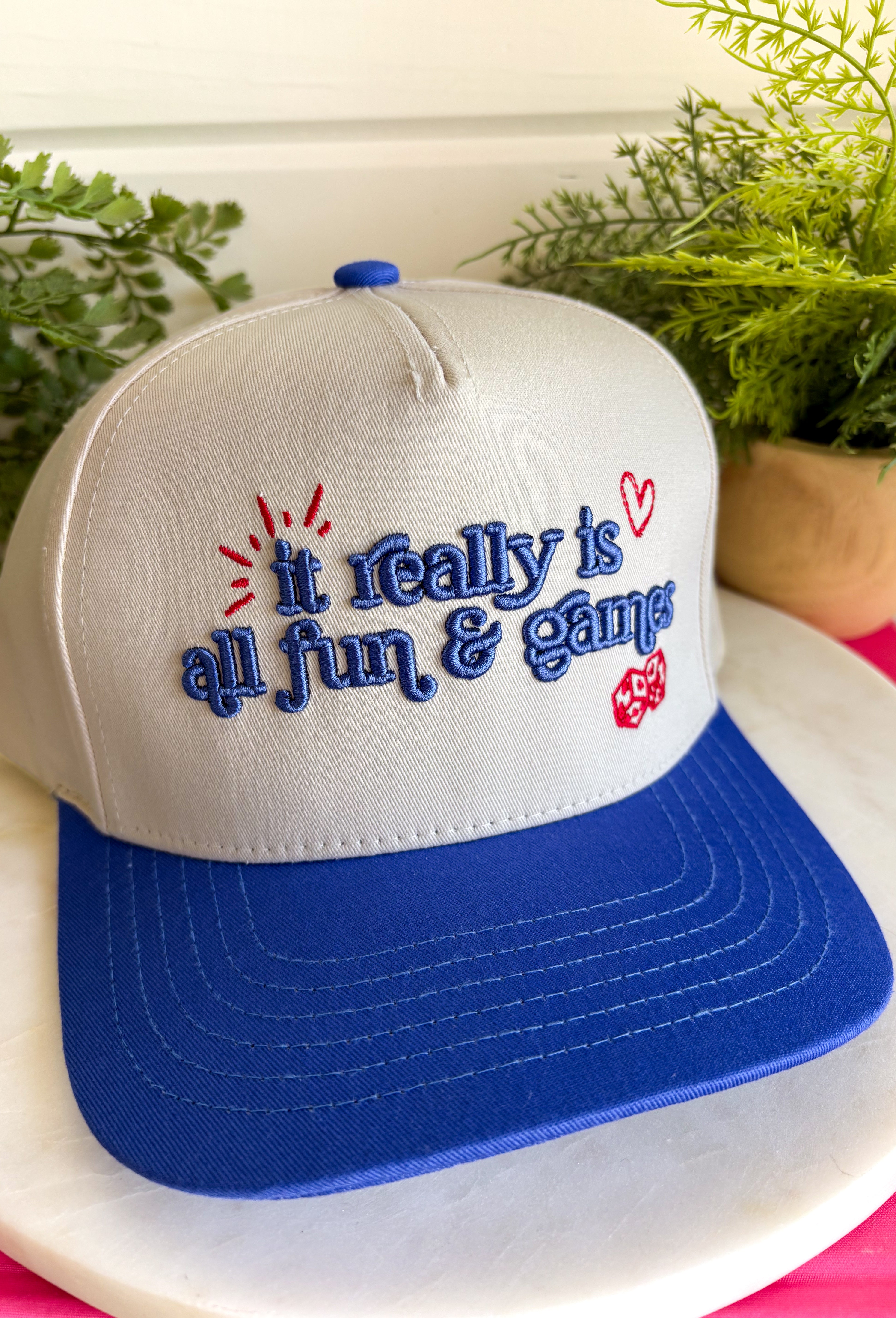 Life's too short not to have a little fun! This trucker hat features&nbsp;"It Really Is All Fun &amp; Games" embroidered in royal blue with red embroidered heart and dice details for the perfect playful touch. The neutral crown paired with the royal blue bill creates a classic color combination that's easy to style, while the fun graphic adds personality to every outfit. 