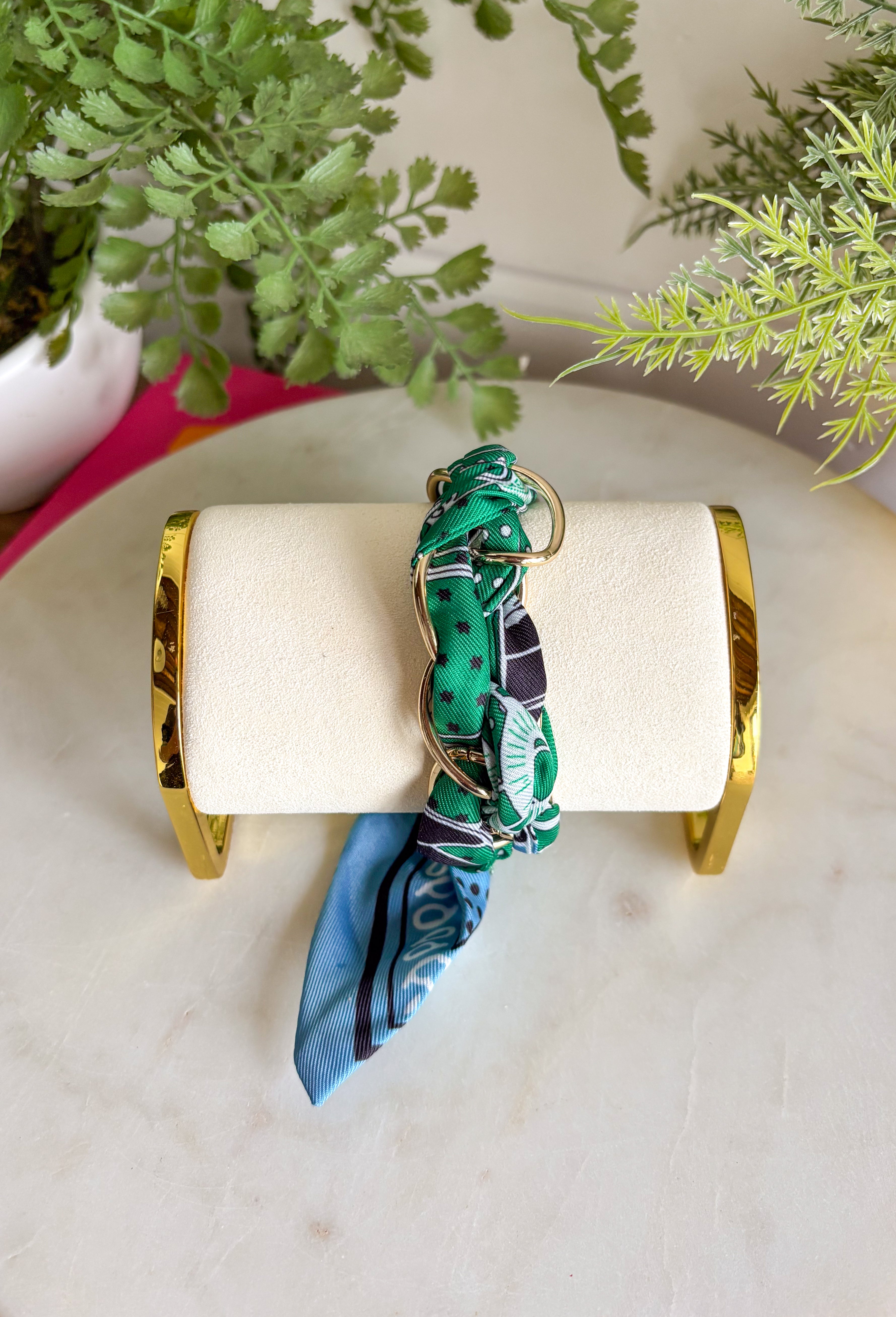 A bold accessory with a cool, laid-back vibe! This fabric wrapped chain link bracelet features oversized gold chain links wrapped in a silky bandana-style fabric. The mix of green and blue tones with pops of black and white creates a fun, eye-catching look that feels both stylish and effortless. The contrast between the polished gold chain and the soft patterned fabric gives this bracelet a unique statement feel. 