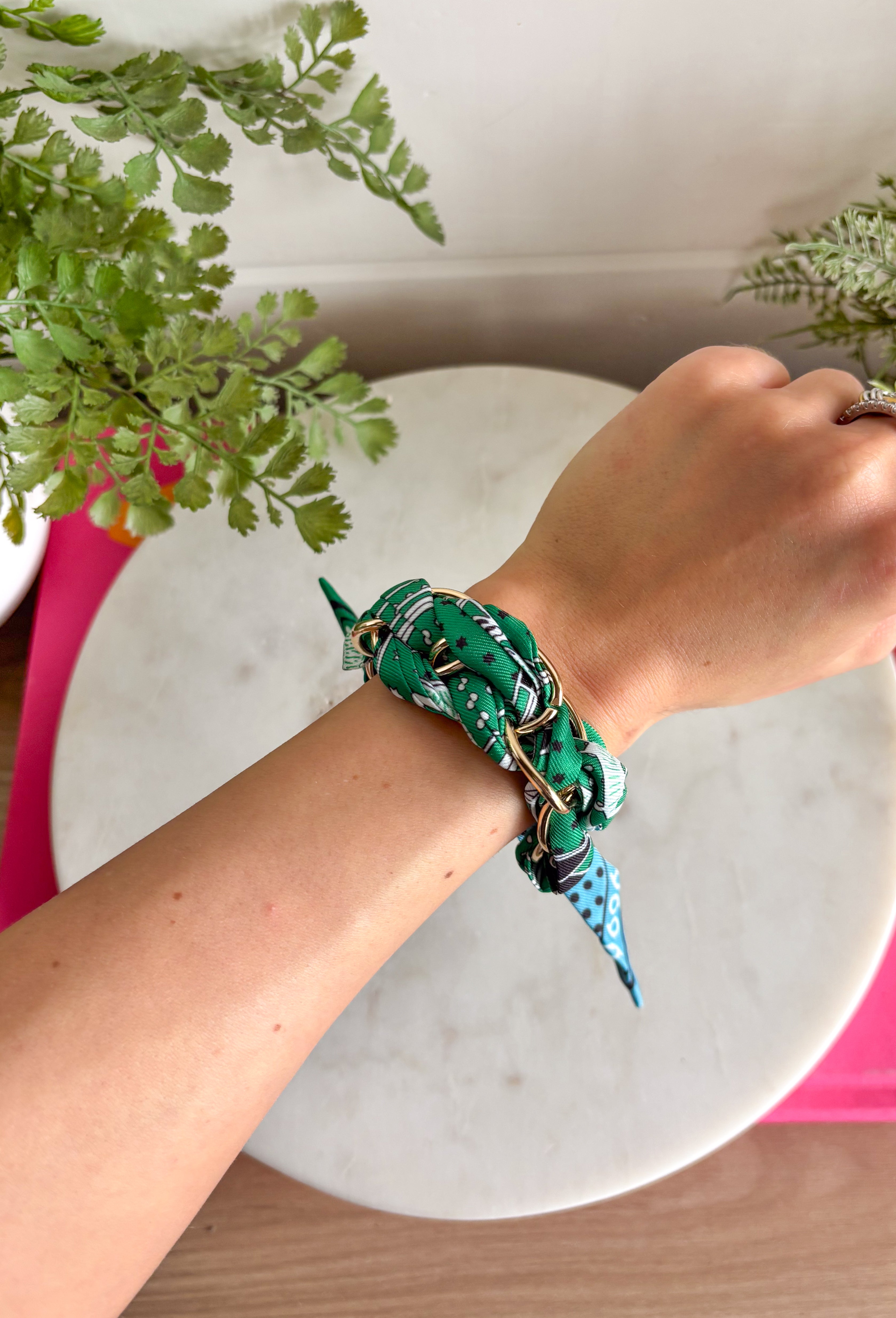 A bold accessory with a cool, laid-back vibe! This fabric wrapped chain link bracelet features oversized gold chain links wrapped in a silky bandana-style fabric. The mix of green and blue tones with pops of black and white creates a fun, eye-catching look that feels both stylish and effortless. The contrast between the polished gold chain and the soft patterned fabric gives this bracelet a unique statement feel. 