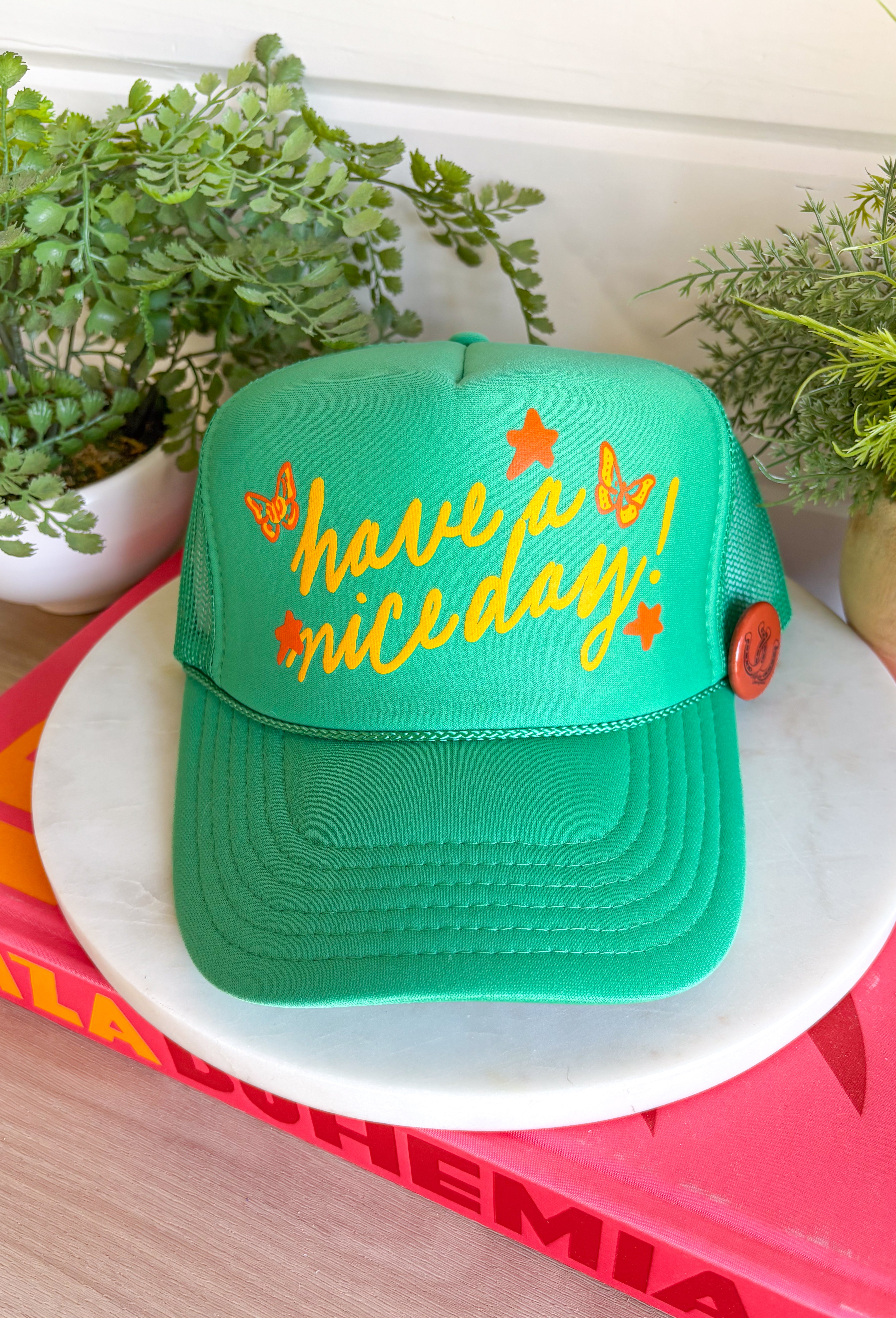 All the good vibes in one hat! This green trucker hat features “Have A Nice Day!” in a warm mustard tone, surrounded by playful orange and mustard butterflies and star details for the happiest little pop of personality. A small rust button on the side with a horse shoe design adds a fun, laid-back touch that makes this hat even more unique. With a breathable mesh backing and adjustable fit, it’s perfect for sunny days, weekend plans, or anytime you're in your carefree era.