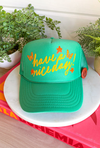 All the good vibes in one hat! This green trucker hat features “Have A Nice Day!” in a warm mustard tone, surrounded by playful orange and mustard butterflies and star details for the happiest little pop of personality. A small rust button on the side with a horse shoe design adds a fun, laid-back touch that makes this hat even more unique. With a breathable mesh backing and adjustable fit, it’s perfect for sunny days, weekend plans, or anytime you're in your carefree era.