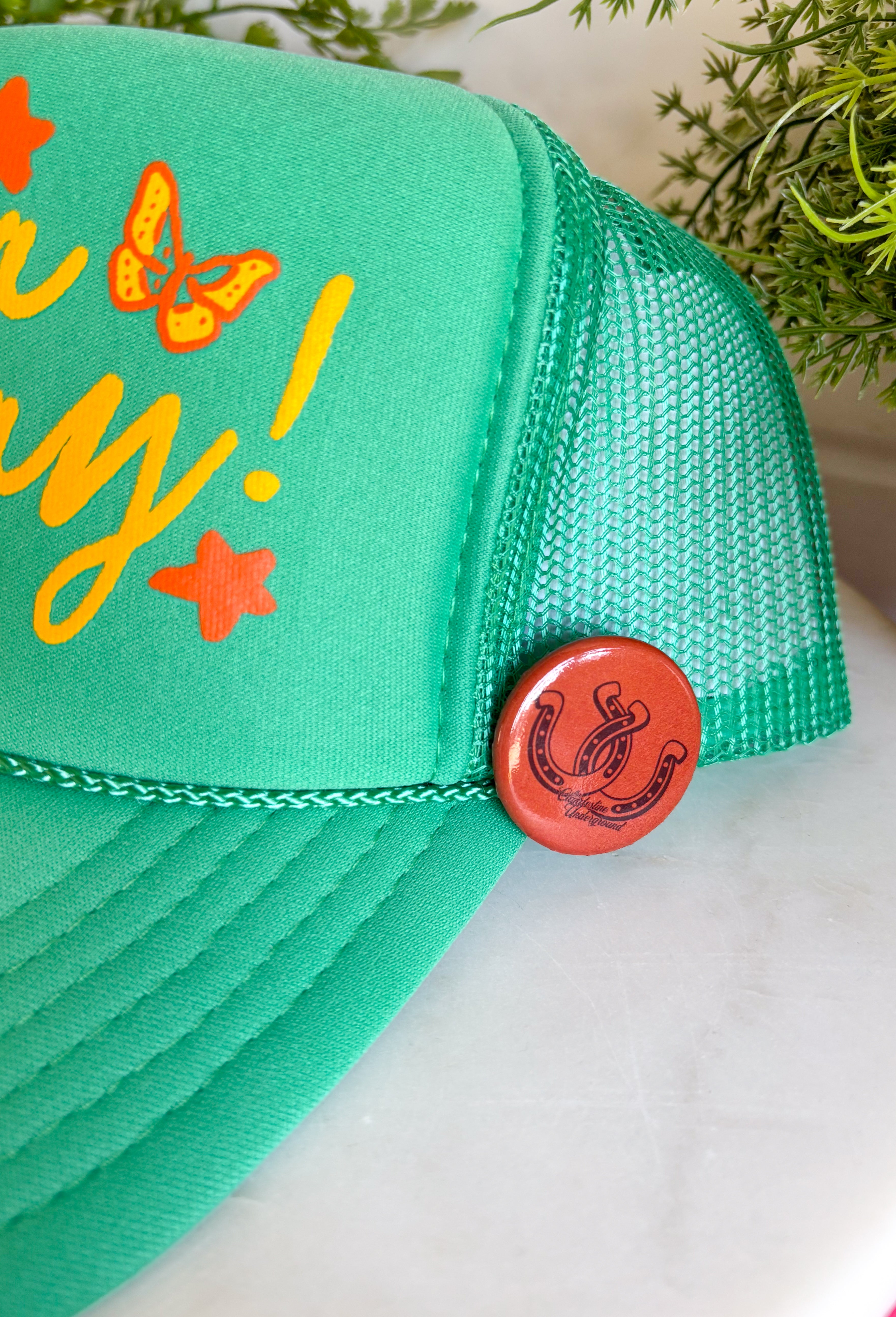 All the good vibes in one hat! This green trucker hat features “Have A Nice Day!” in a warm mustard tone, surrounded by playful orange and mustard butterflies and star details for the happiest little pop of personality. A small rust button on the side with a horse shoe design adds a fun, laid-back touch that makes this hat even more unique. With a breathable mesh backing and adjustable fit, it’s perfect for sunny days, weekend plans, or anytime you're in your carefree era.