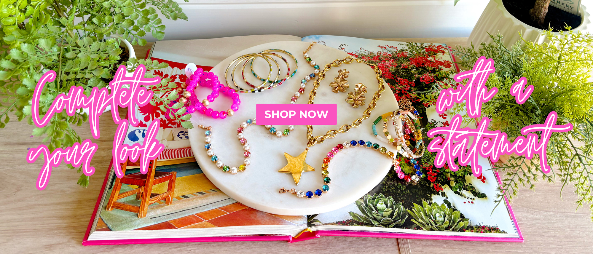 "complete your look with a statement" "shop now" colorful jewelry displayed on a marble slab layered on an open book surrounded by green plants 
