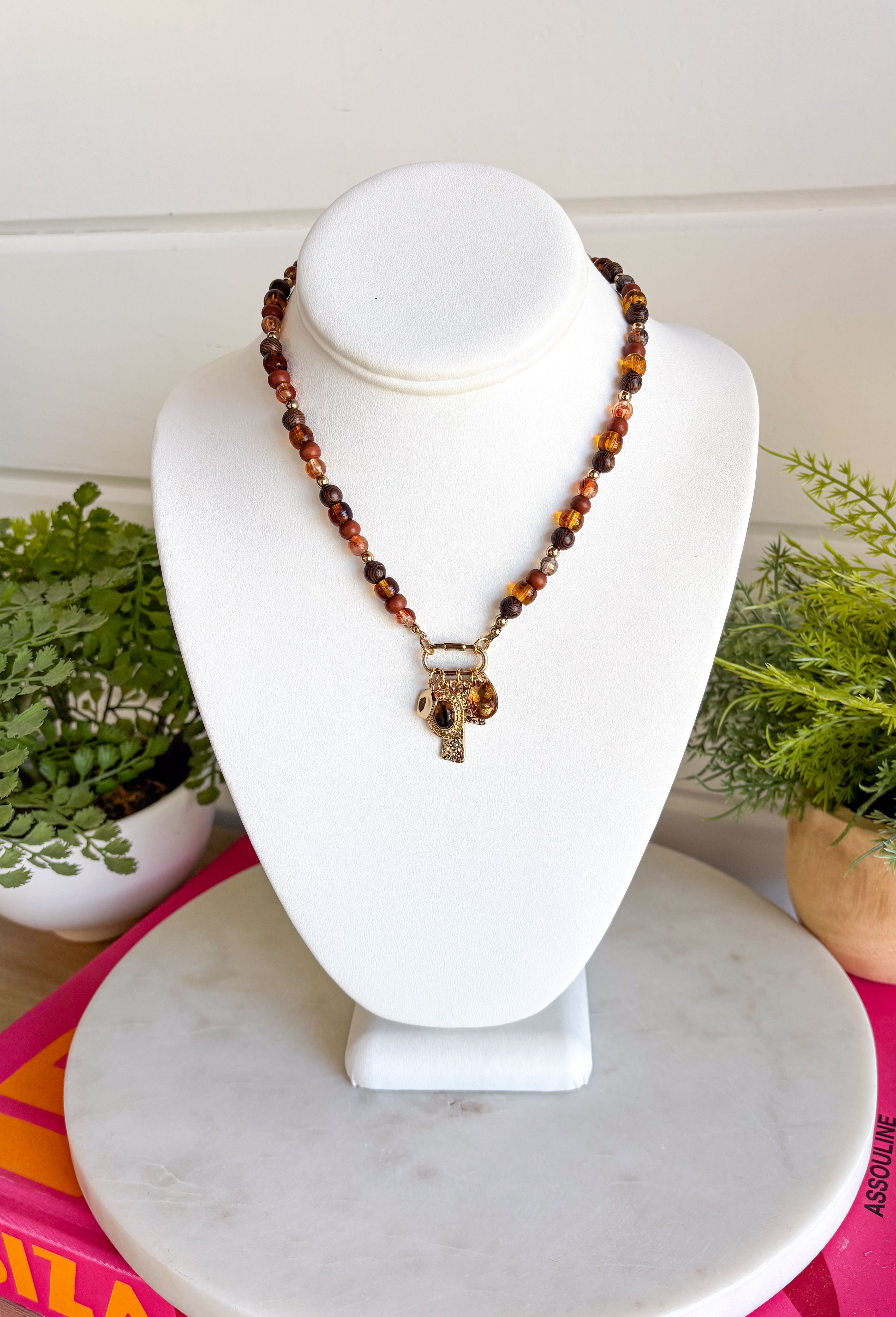Rich earthy tones and meaningful details come together beautifully in this charm necklace that's perfect for the fall season. Featuring warm brown beads, this eye-catching piece is adorned with a mix of charms, including a gold cross, a natural brown stone, a sparkling brown crystal, and hammered gold accents for added texture and shine. Whether worn with a cozy sweater or your favorite dress, this unique necklace adds effortless charm and personality to every outfit.