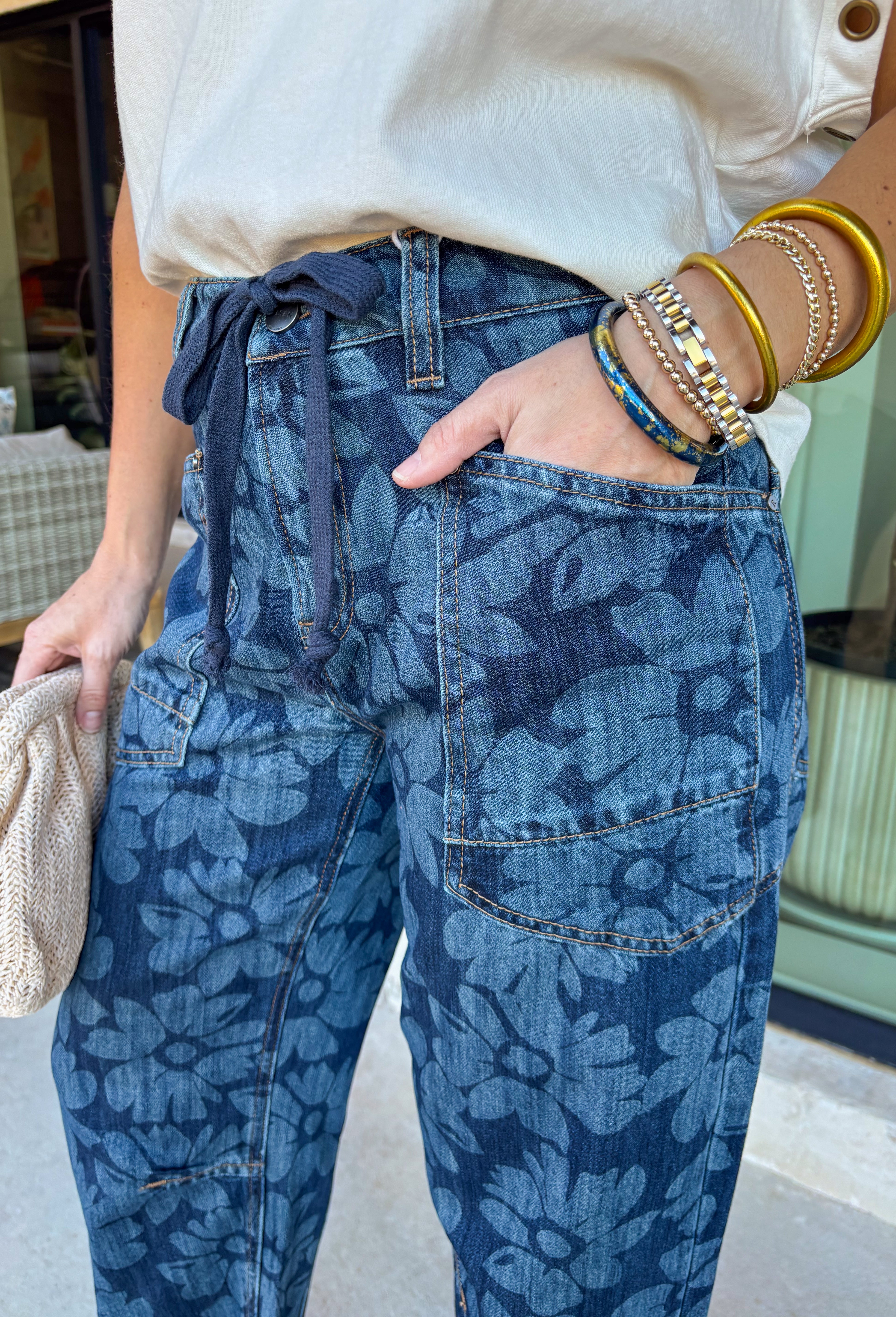 These dark wash barrel jeans bring a fresh, modern twist to everyday denim with a subtle monochromatic floral print woven throughout. The medium wash floral design adds just enough texture and interest while keeping the look sleek and easy to style. Finished with a tie detail at the waist, these jeans offer an adjustable, comfortable fit that still feels polished. The barrel-leg silhouette gives them that relaxed, on-trend shape that pairs perfectly with everything from fitted tees to flowy spring tops.