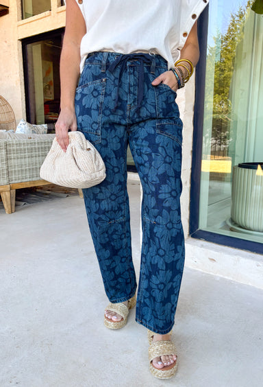 These dark wash barrel jeans bring a fresh, modern twist to everyday denim with a subtle monochromatic floral print woven throughout. The medium wash floral design adds just enough texture and interest while keeping the look sleek and easy to style. Finished with a tie detail at the waist, these jeans offer an adjustable, comfortable fit that still feels polished. The barrel-leg silhouette gives them that relaxed, on-trend shape that pairs perfectly with everything from fitted tees to flowy spring tops.