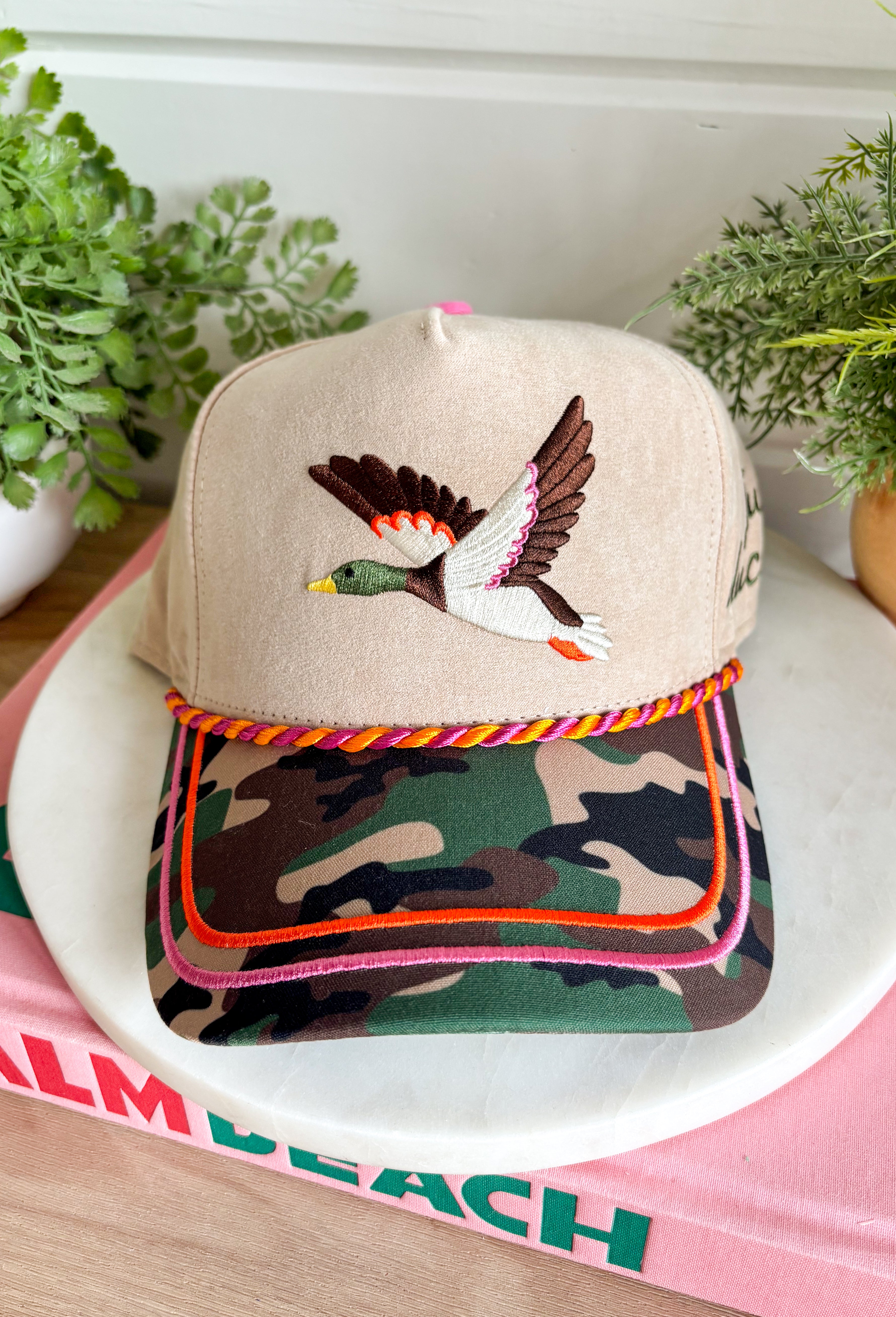 Add a playful outdoor-inspired touch to your everyday look with this unique trucker hat. Featuring a neutral faux suede front and a camouflage bill, this standout style is embroidered with a detailed mallard design accented by pink and orange stitching for an unexpected pop of color. The side is embroidered with "Just Ducky," while the back features "It's A Good Life" in olive green, giving this hat personality from every angle. 