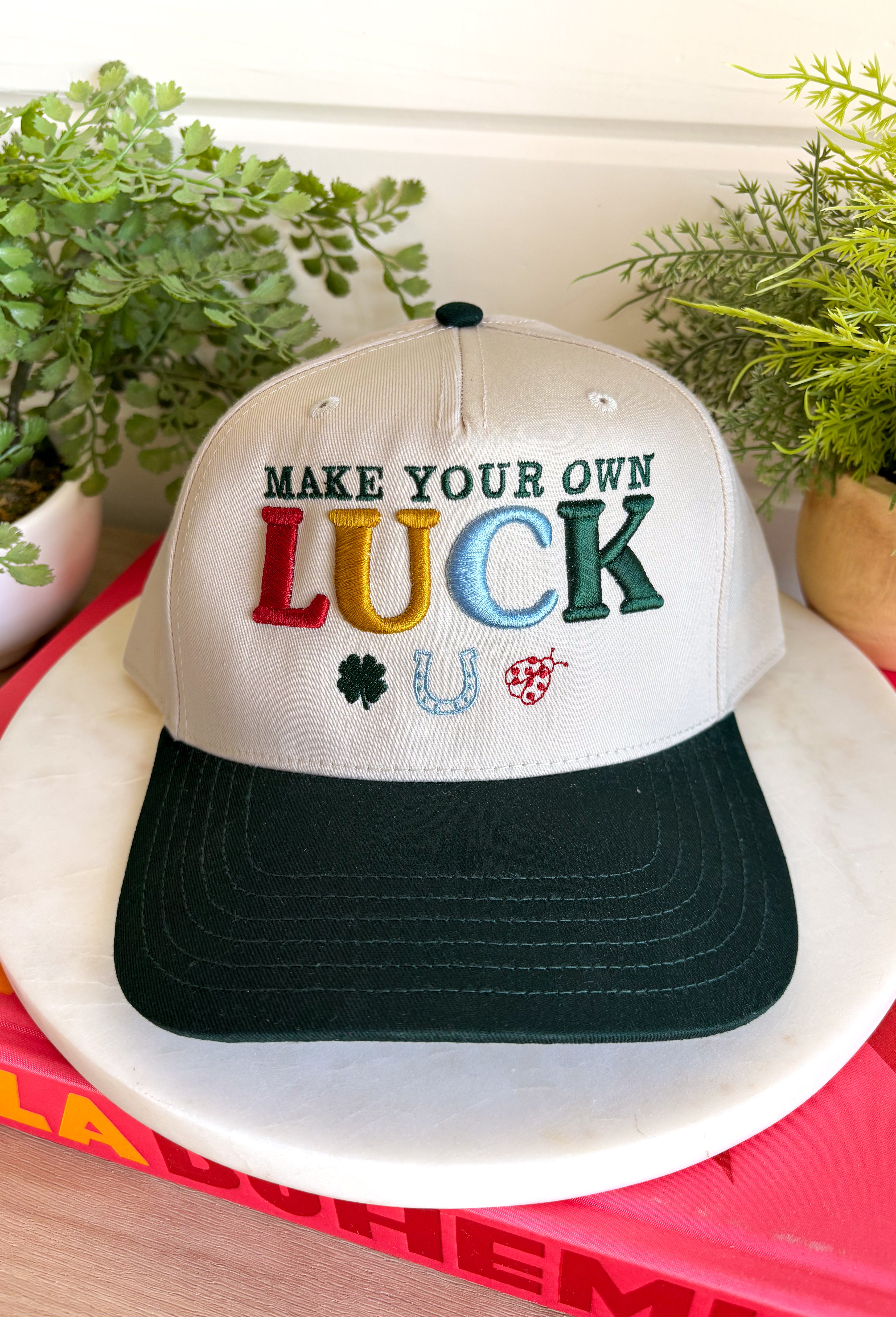A little luck and a lot of style! This trucker hat features beautifully embroidered "Make Your Own Luck" lettering alongside a four-leaf clover, horseshoe, and ladybug in rich, eye-catching colors. The neutral crown paired with the dark green bill gives it a timeless look that's easy to style, while the detailed embroidery adds the perfect touch of personality. 
