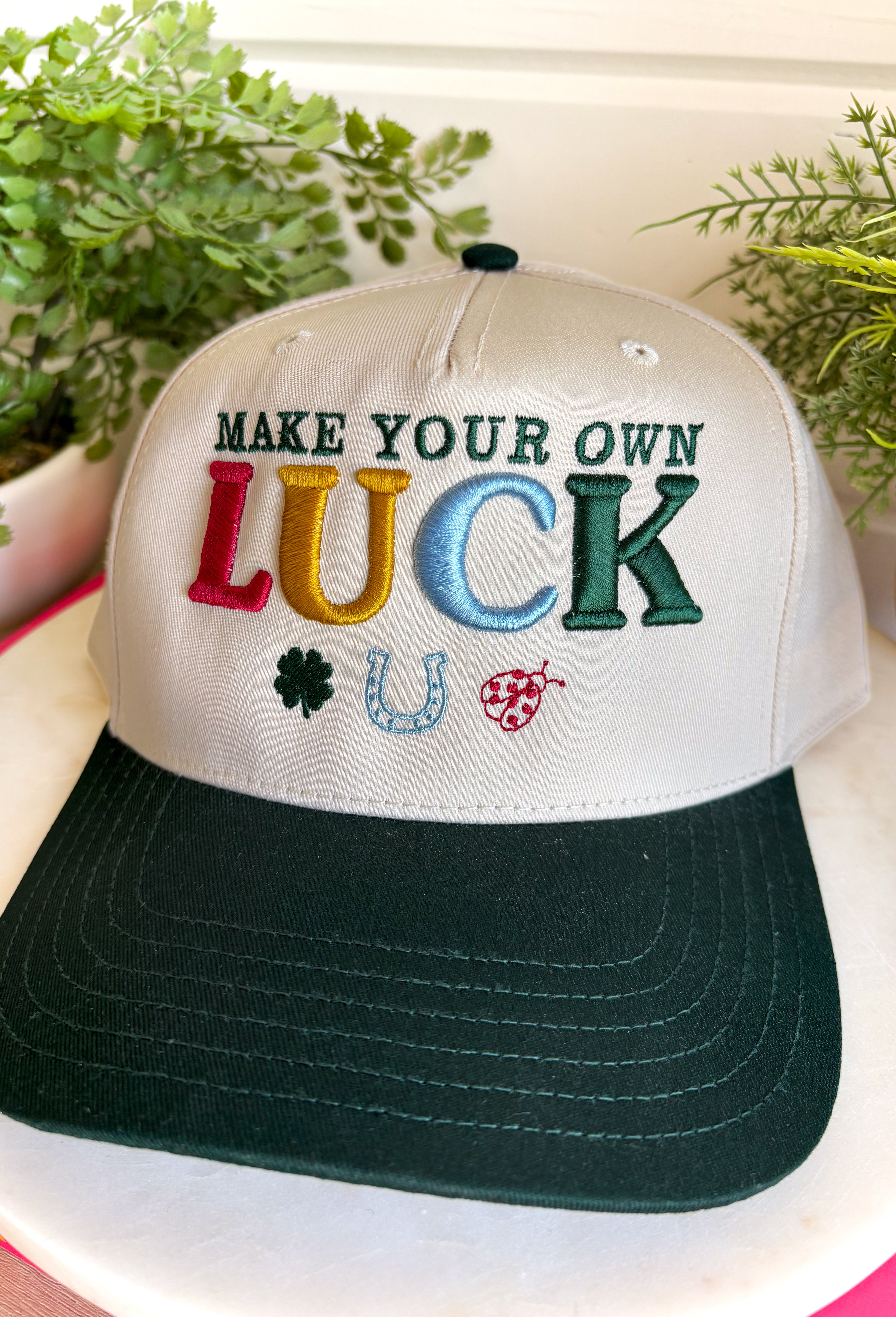 A little luck and a lot of style! This trucker hat features beautifully embroidered "Make Your Own Luck" lettering alongside a four-leaf clover, horseshoe, and ladybug in rich, eye-catching colors. The neutral crown paired with the dark green bill gives it a timeless look that's easy to style, while the detailed embroidery adds the perfect touch of personality. 