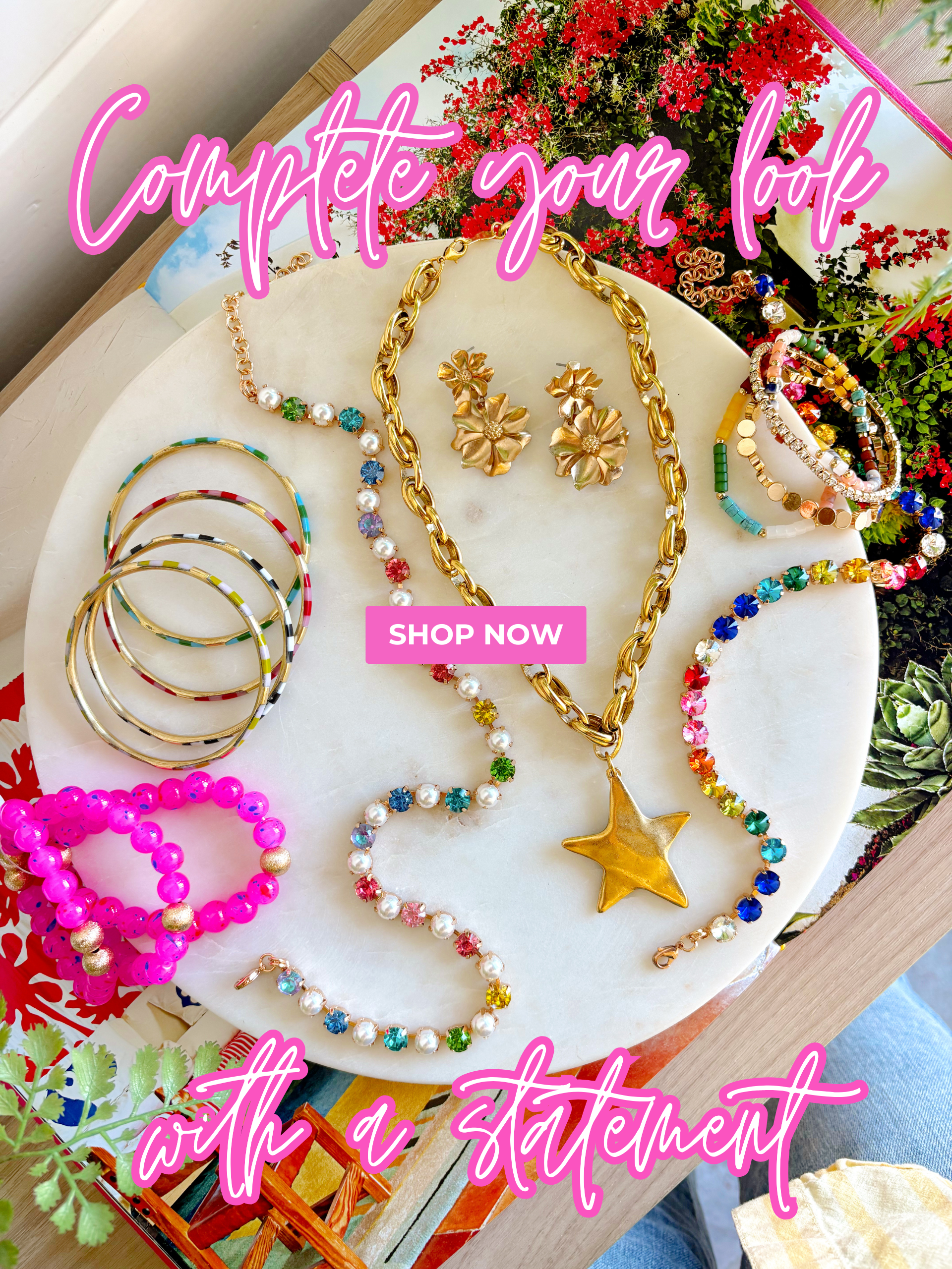 "complete your look with a statement" "shop now" colorful jewelry displayed on a marble slab layered on an open book surrounded by green plants 