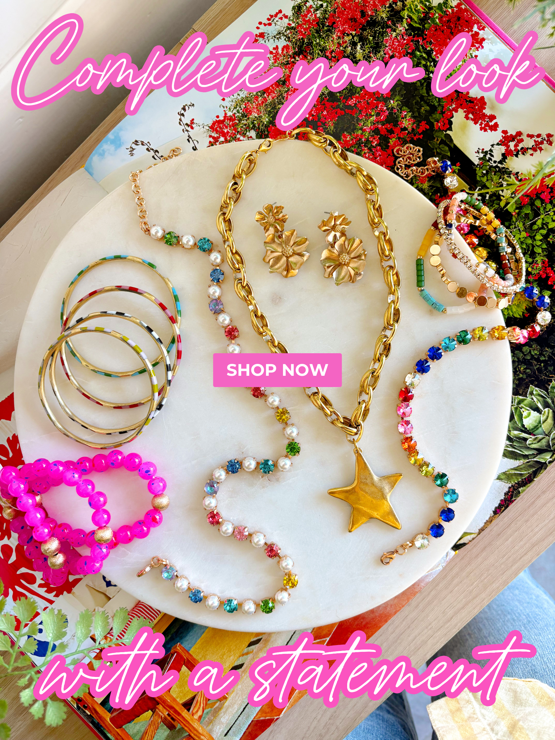 "complete your look with a statement" "shop now" colorful jewelry displayed on a marble slab layered on an open book surrounded by green plants 