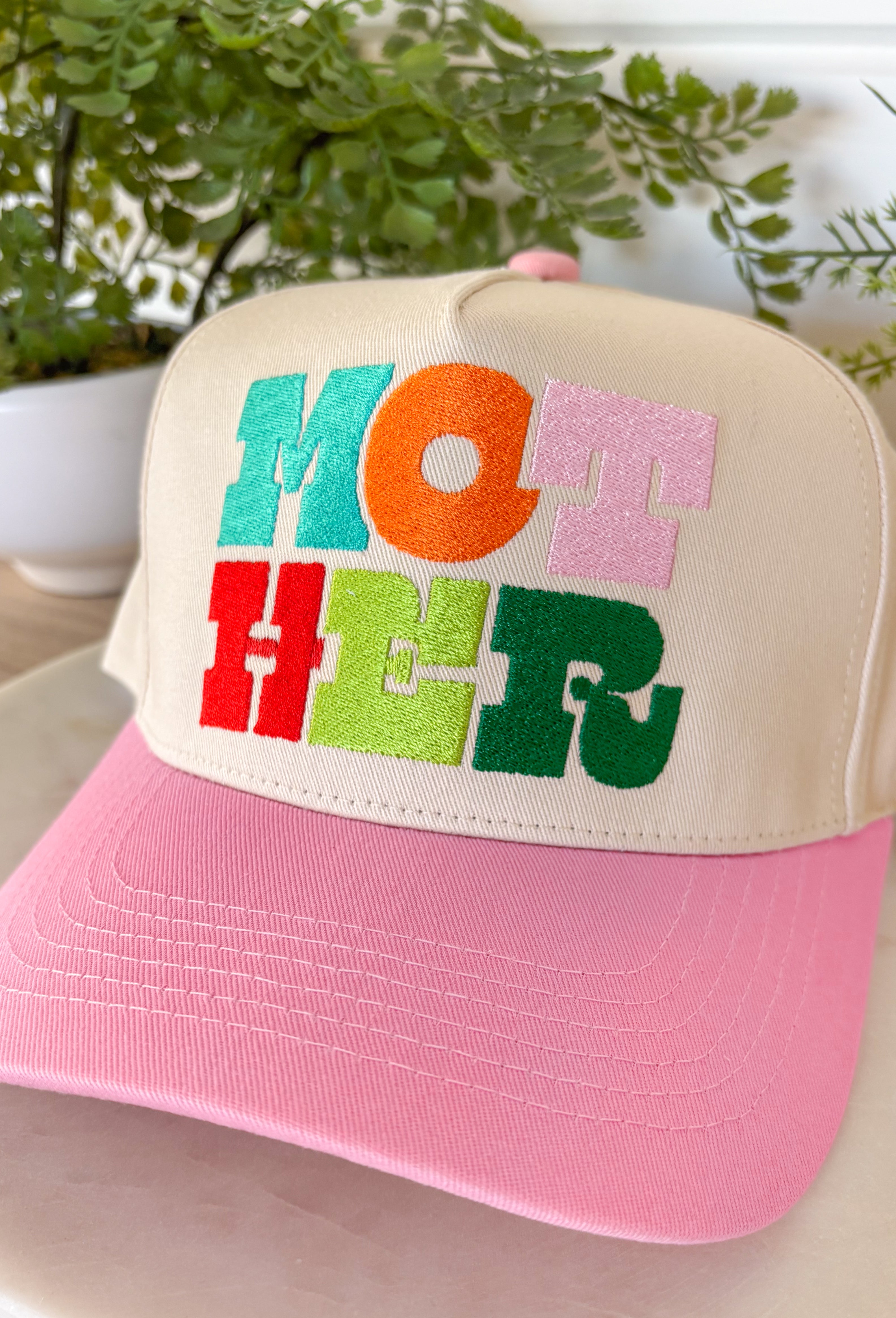This neutral trucker hat is a sweet, feel-good accessory with a fun pop of color. Designed with a soft light pink bill and “Mother” embroidered across the front in a colorful print, it’s playful, meaningful, and easy to wear. The neutral base keeps it versatile, while the adjustable back ensures a comfortable, customizable fit. Perfect for everyday errands, casual outings, or gifting to a mama who deserves a little extra love.