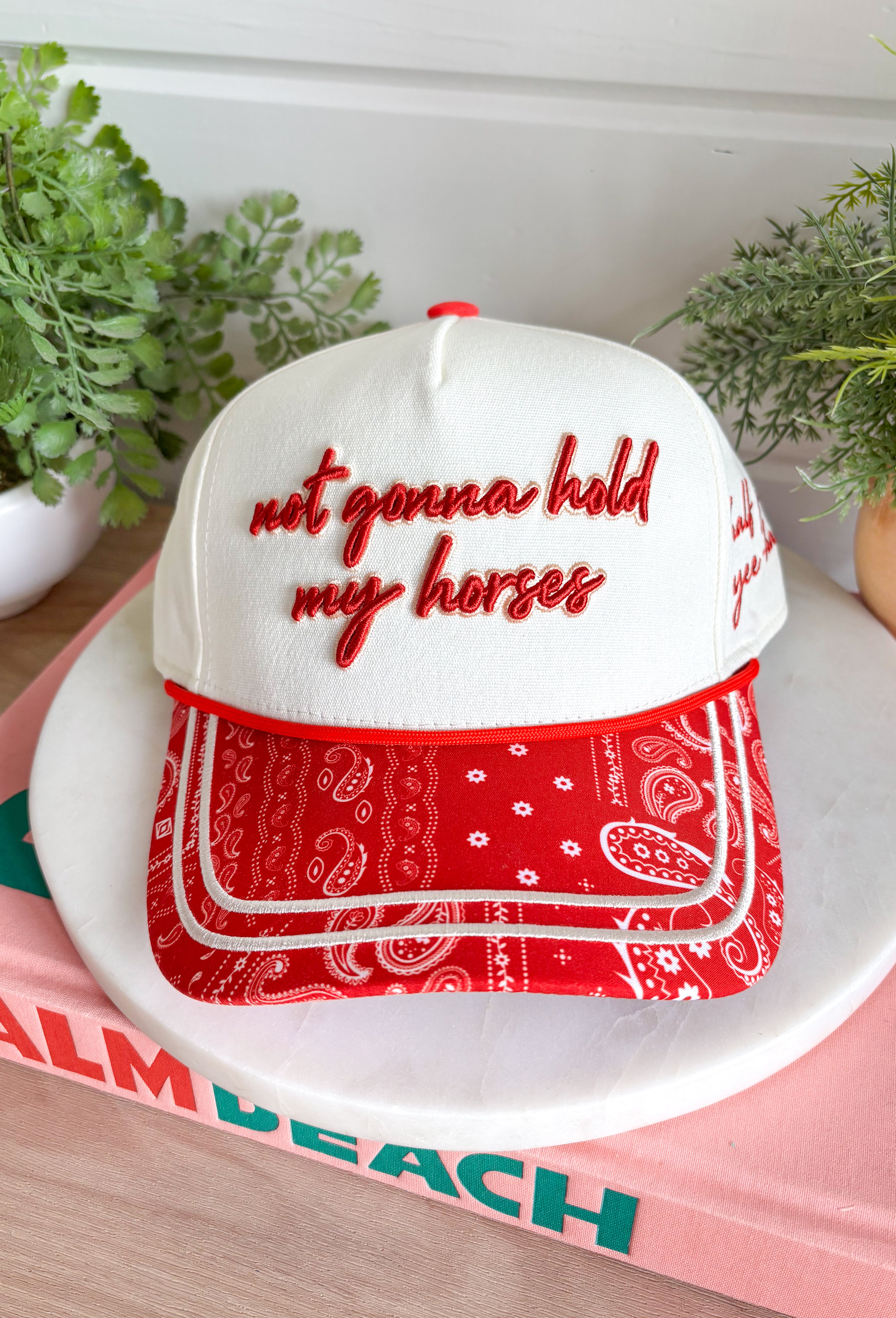 Bring a little western charm and plenty of personality to your everyday style with this fun trucker hat. Featuring a neutral front paired with a bold red bandana print bill, this standout accessory is embroidered with "Not Gonna Hold My Horses" across the front in vibrant red stitching. The side is finished with "Half Past Yee-Haw" embroidery, while a red embroidered horseshoe on the back adds the perfect finishing touch. 