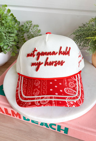 Bring a little western charm and plenty of personality to your everyday style with this fun trucker hat. Featuring a neutral front paired with a bold red bandana print bill, this standout accessory is embroidered with "Not Gonna Hold My Horses" across the front in vibrant red stitching. The side is finished with "Half Past Yee-Haw" embroidery, while a red embroidered horseshoe on the back adds the perfect finishing touch. 