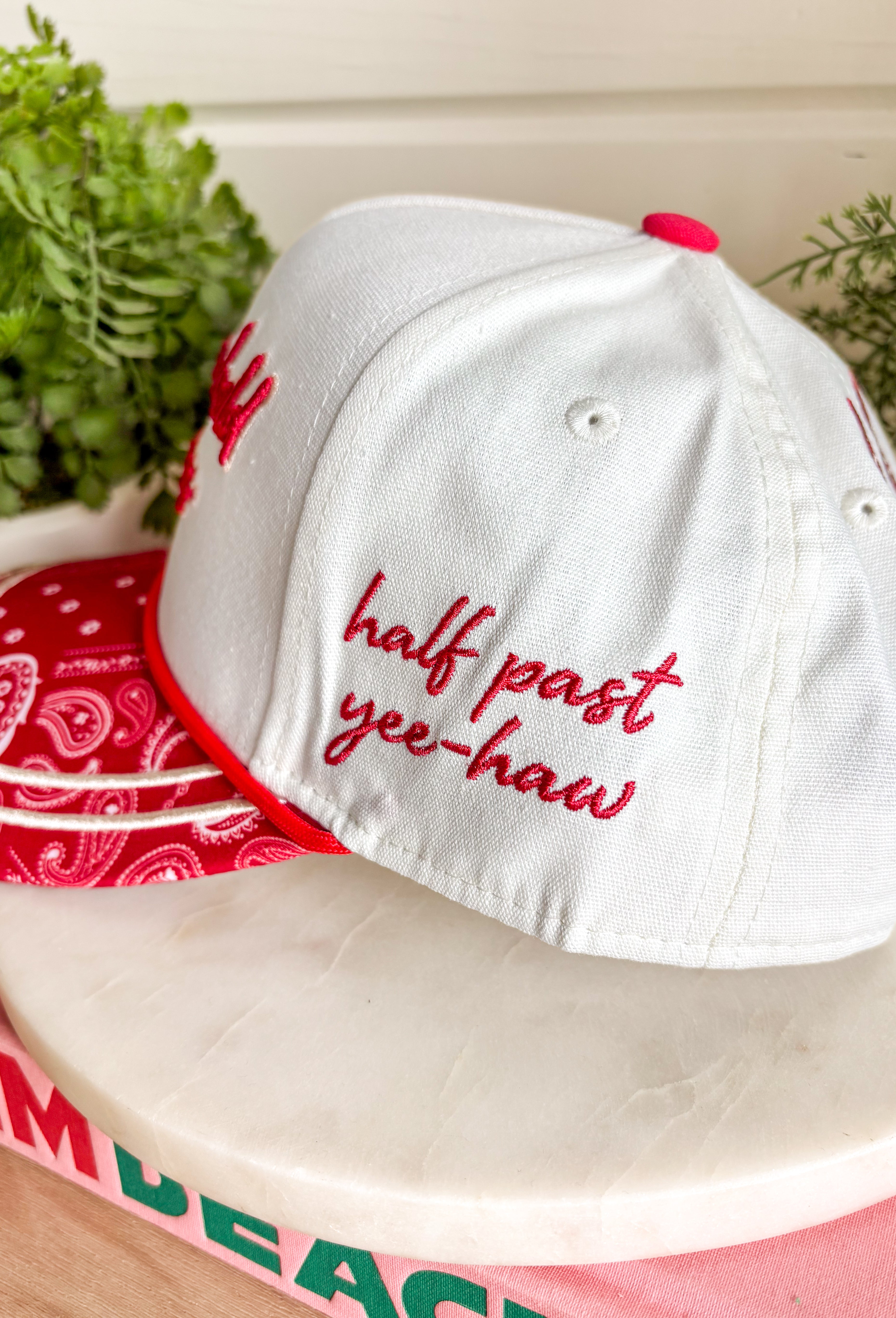 Bring a little western charm and plenty of personality to your everyday style with this fun trucker hat. Featuring a neutral front paired with a bold red bandana print bill, this standout accessory is embroidered with "Not Gonna Hold My Horses" across the front in vibrant red stitching. The side is finished with "Half Past Yee-Haw" embroidery, while a red embroidered horseshoe on the back adds the perfect finishing touch. 