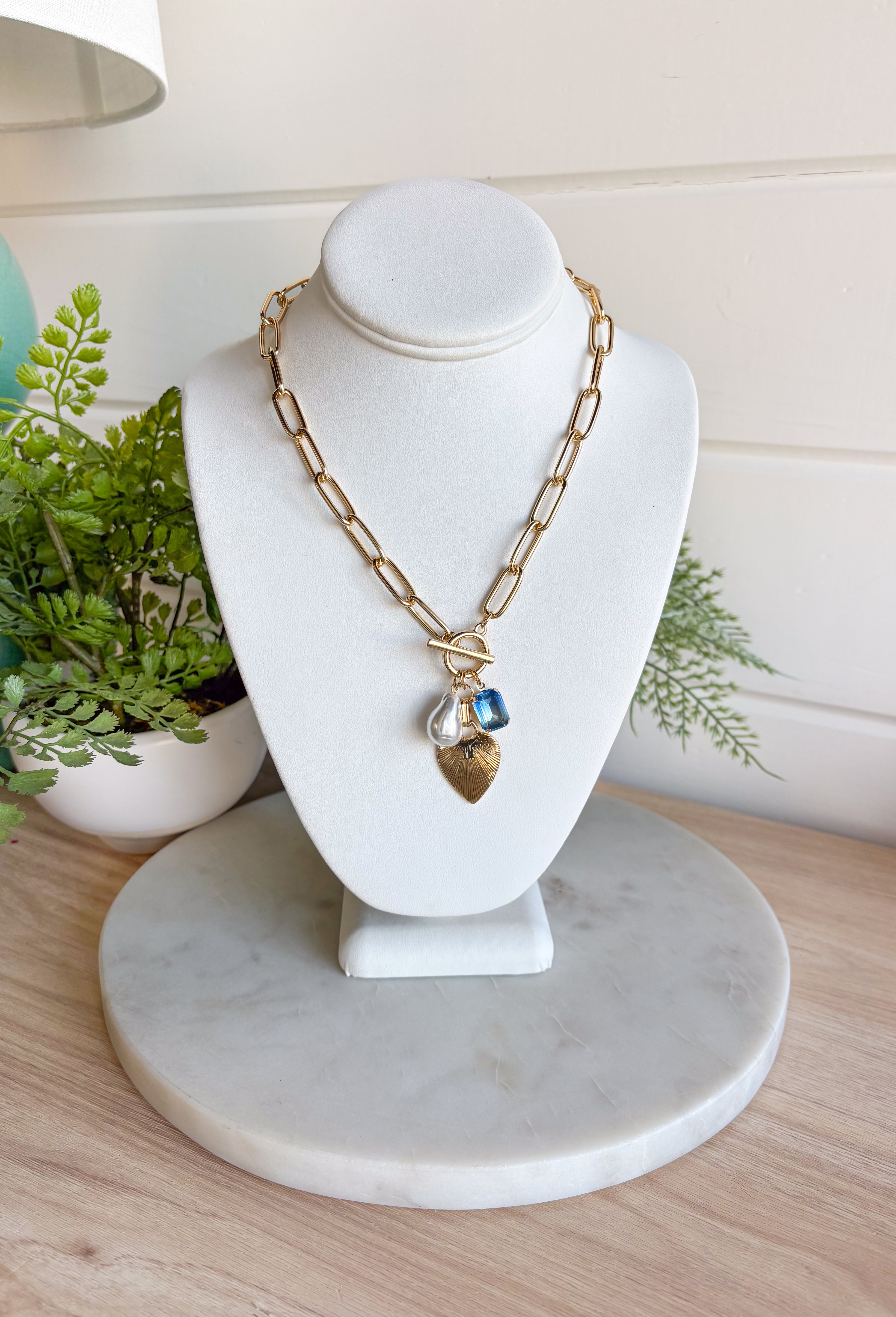A little bit classic, a little bit playful — this gold paperclip chain necklace is the perfect piece to wear all year long. Designed with a mix of charming details like a textured heart, a pearl accent, and a touch of blue rhinestone sparkle, it adds just the right amount of personality to any look. Finished with a toggle clasp for easy wear, it’s made to shine from everyday outfits to special occasions.