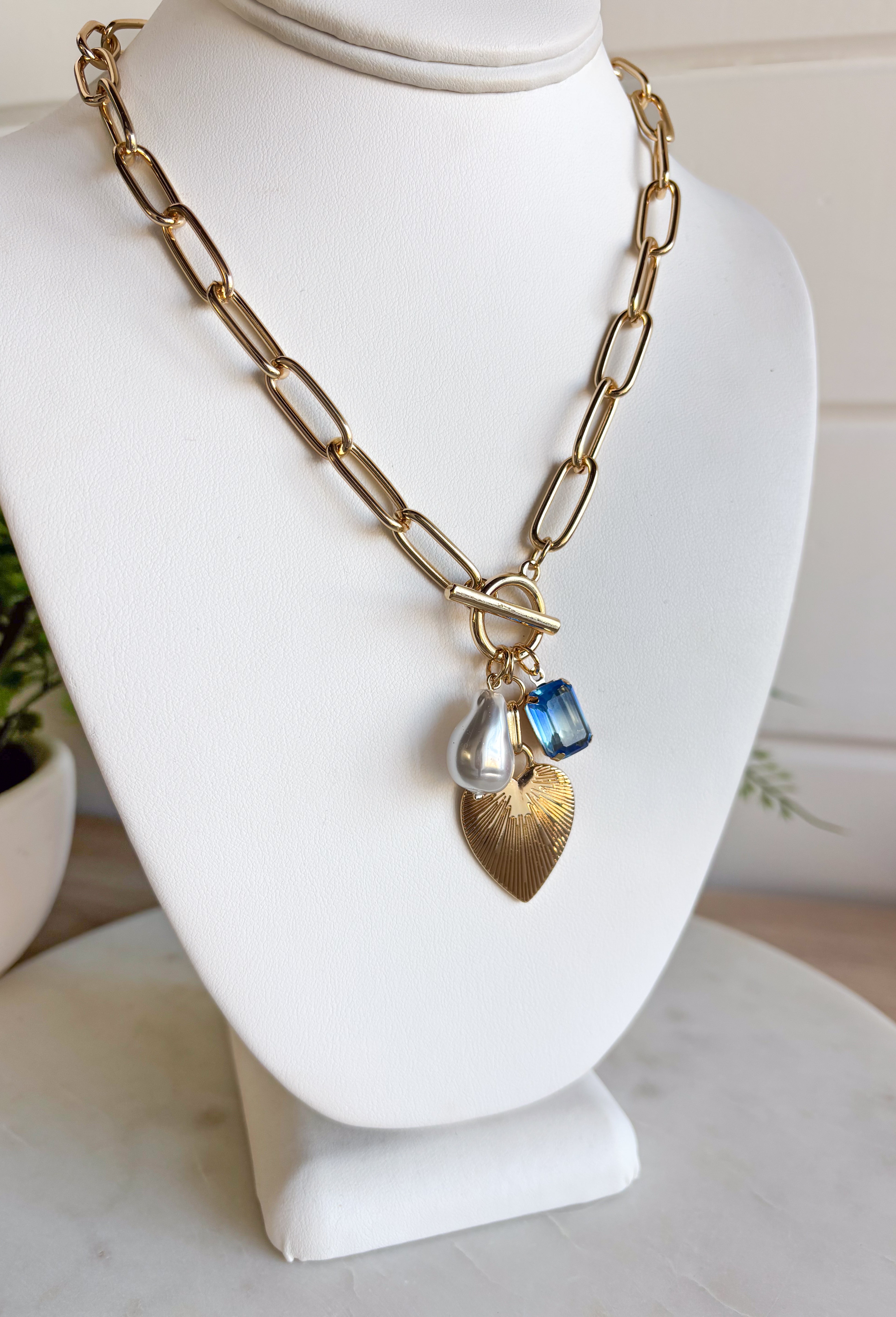 A little bit classic, a little bit playful — this gold paperclip chain necklace is the perfect piece to wear all year long. Designed with a mix of charming details like a textured heart, a pearl accent, and a touch of blue rhinestone sparkle, it adds just the right amount of personality to any look. Finished with a toggle clasp for easy wear, it’s made to shine from everyday outfits to special occasions.