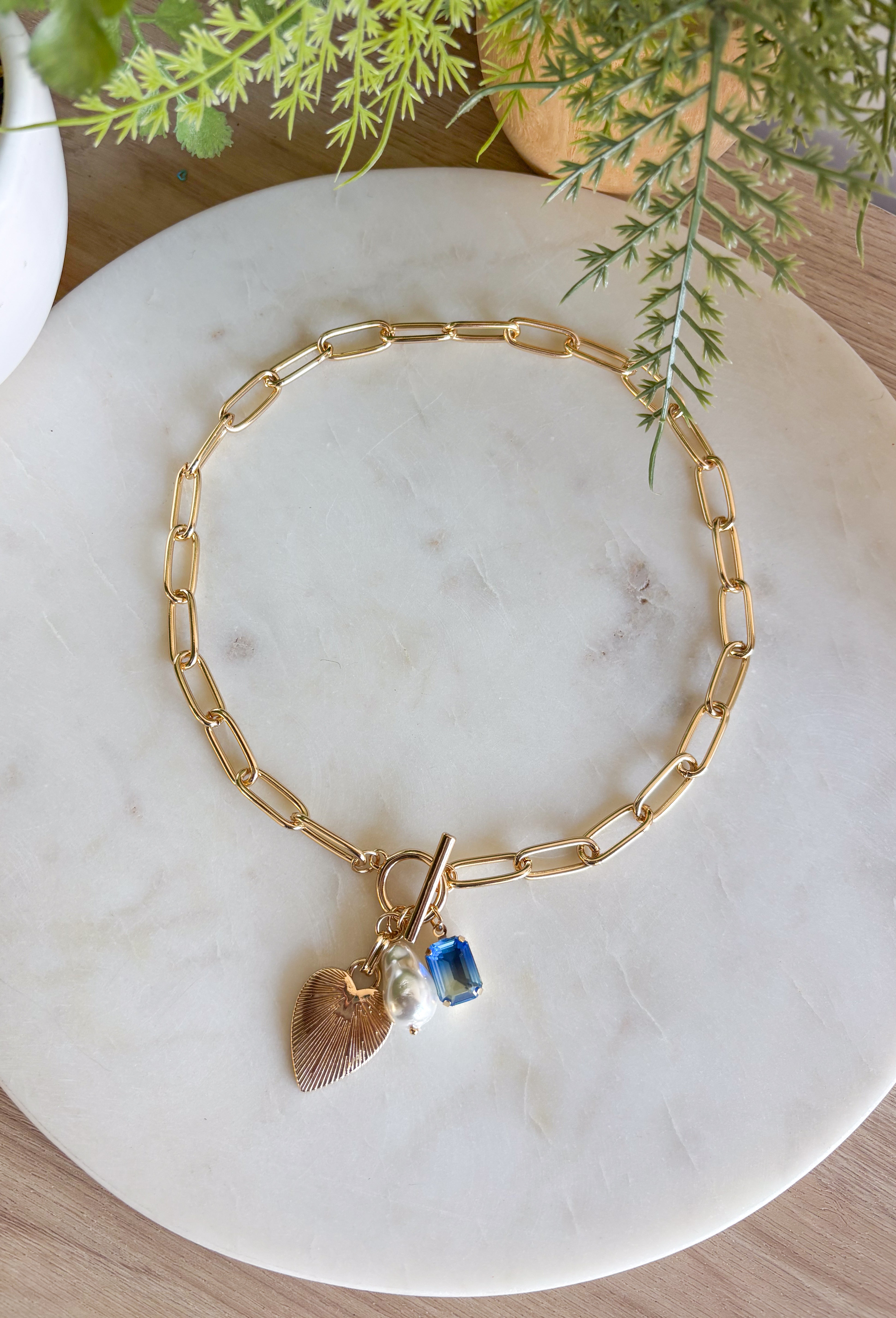 A little bit classic, a little bit playful — this gold paperclip chain necklace is the perfect piece to wear all year long. Designed with a mix of charming details like a textured heart, a pearl accent, and a touch of blue rhinestone sparkle, it adds just the right amount of personality to any look. Finished with a toggle clasp for easy wear, it’s made to shine from everyday outfits to special occasions.