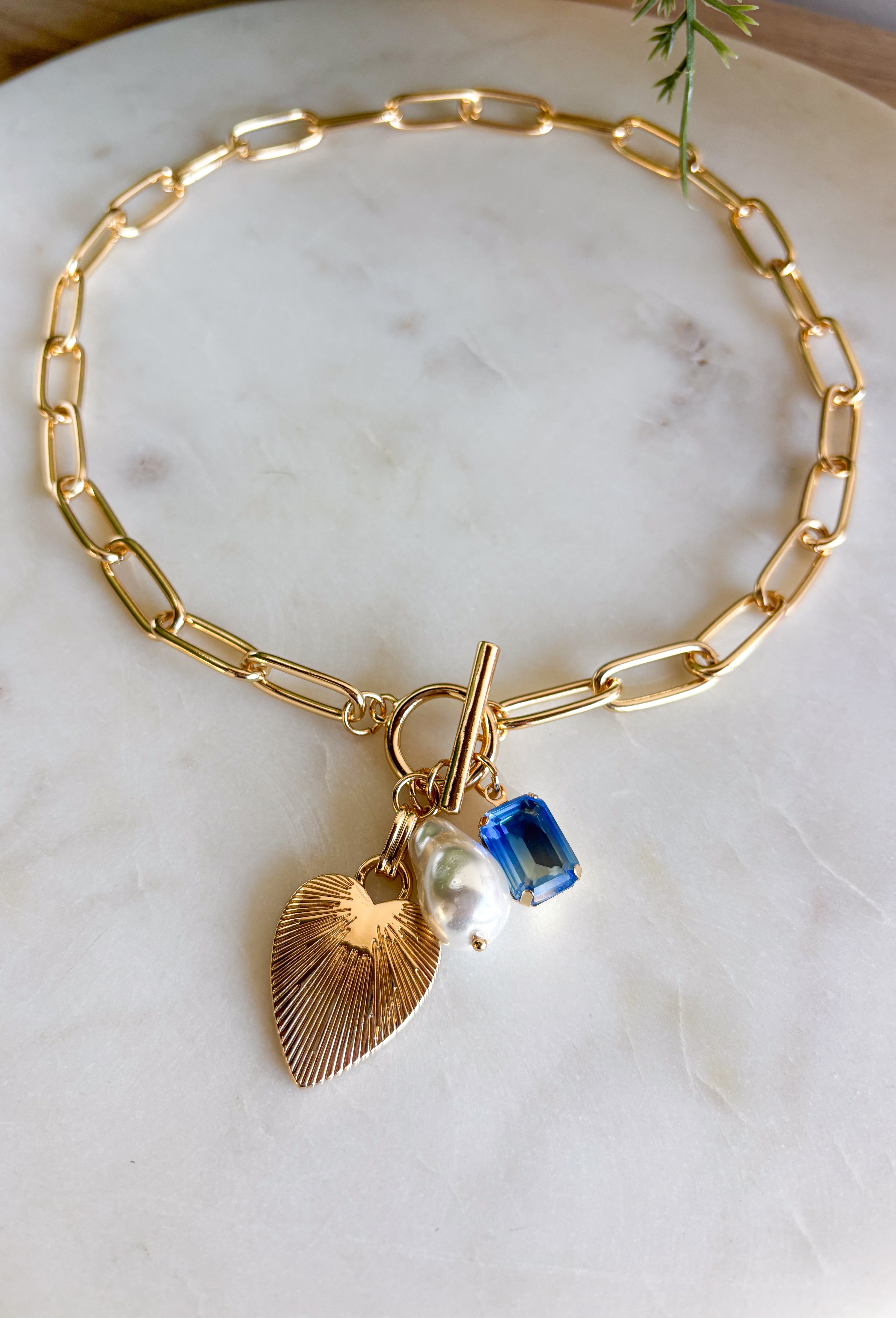 A little bit classic, a little bit playful — this gold paperclip chain necklace is the perfect piece to wear all year long. Designed with a mix of charming details like a textured heart, a pearl accent, and a touch of blue rhinestone sparkle, it adds just the right amount of personality to any look. Finished with a toggle clasp for easy wear, it’s made to shine from everyday outfits to special occasions.