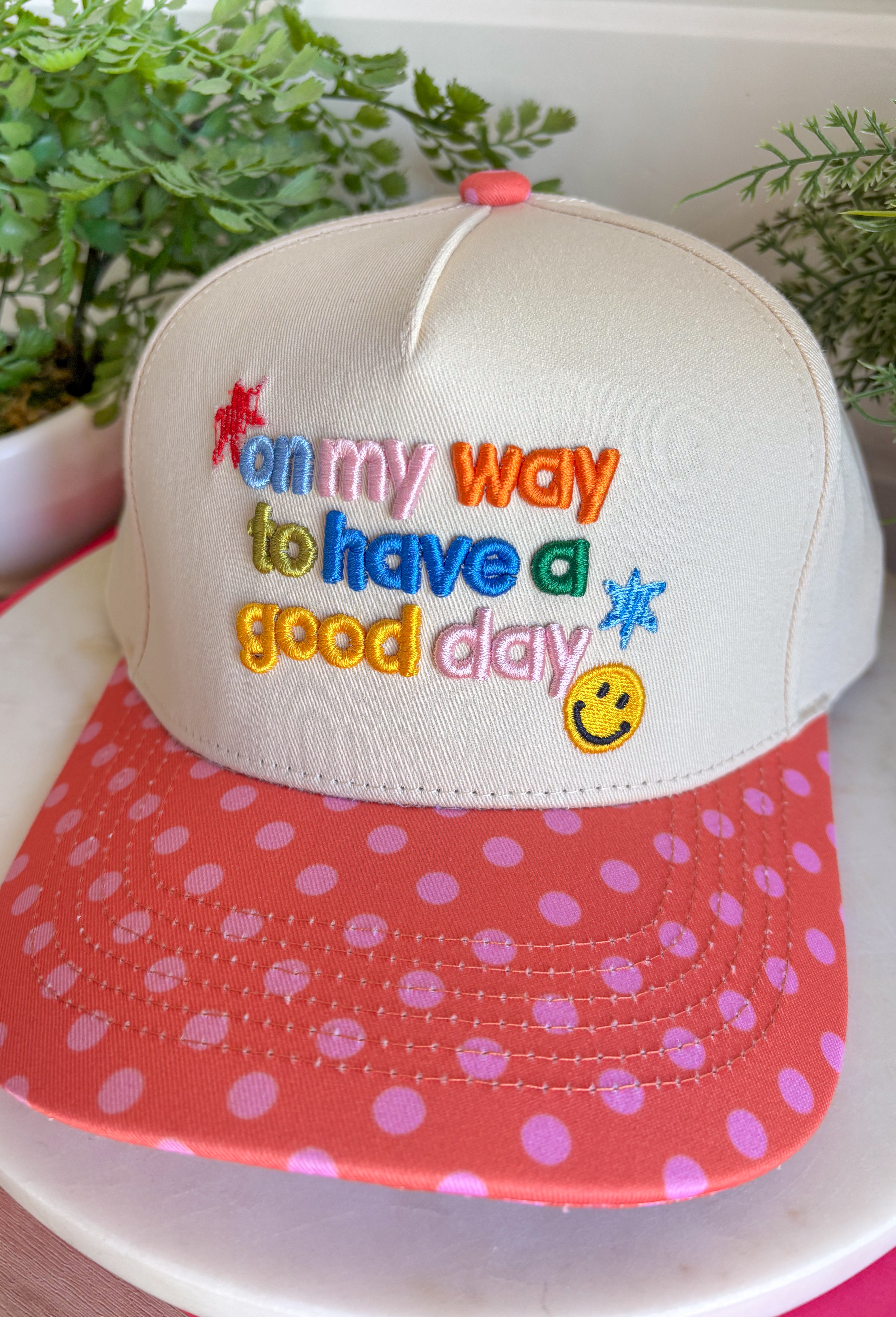 Start every day with a smile in this cheerful trucker hat. Featuring "On My Way To Have A Good Day" embroidered in colorful thread, it's the perfect reminder to embrace every adventure with a positive attitude. The coral bill with playful pink polka dots and matching top button add a fun pop of color, making this hat a standout accessory for everything from coffee runs to weekend road trips. Finished with an adjustable fit, it's the perfect blend of comfort, personality, and everyday style.