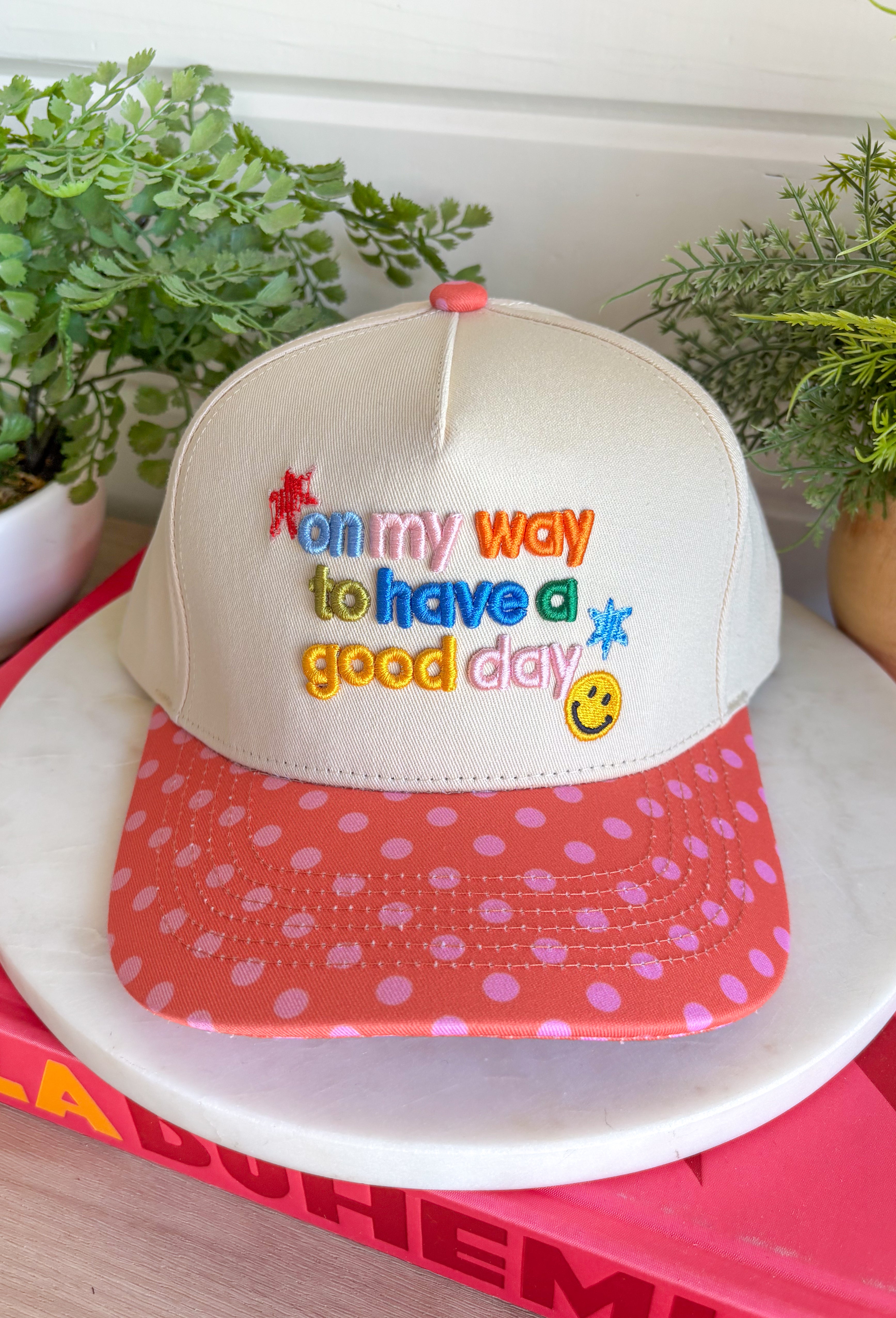 Start every day with a smile in this cheerful trucker hat. Featuring "On My Way To Have A Good Day" embroidered in colorful thread, it's the perfect reminder to embrace every adventure with a positive attitude. The coral bill with playful pink polka dots and matching top button add a fun pop of color, making this hat a standout accessory for everything from coffee runs to weekend road trips. Finished with an adjustable fit, it's the perfect blend of comfort, personality, and everyday style.