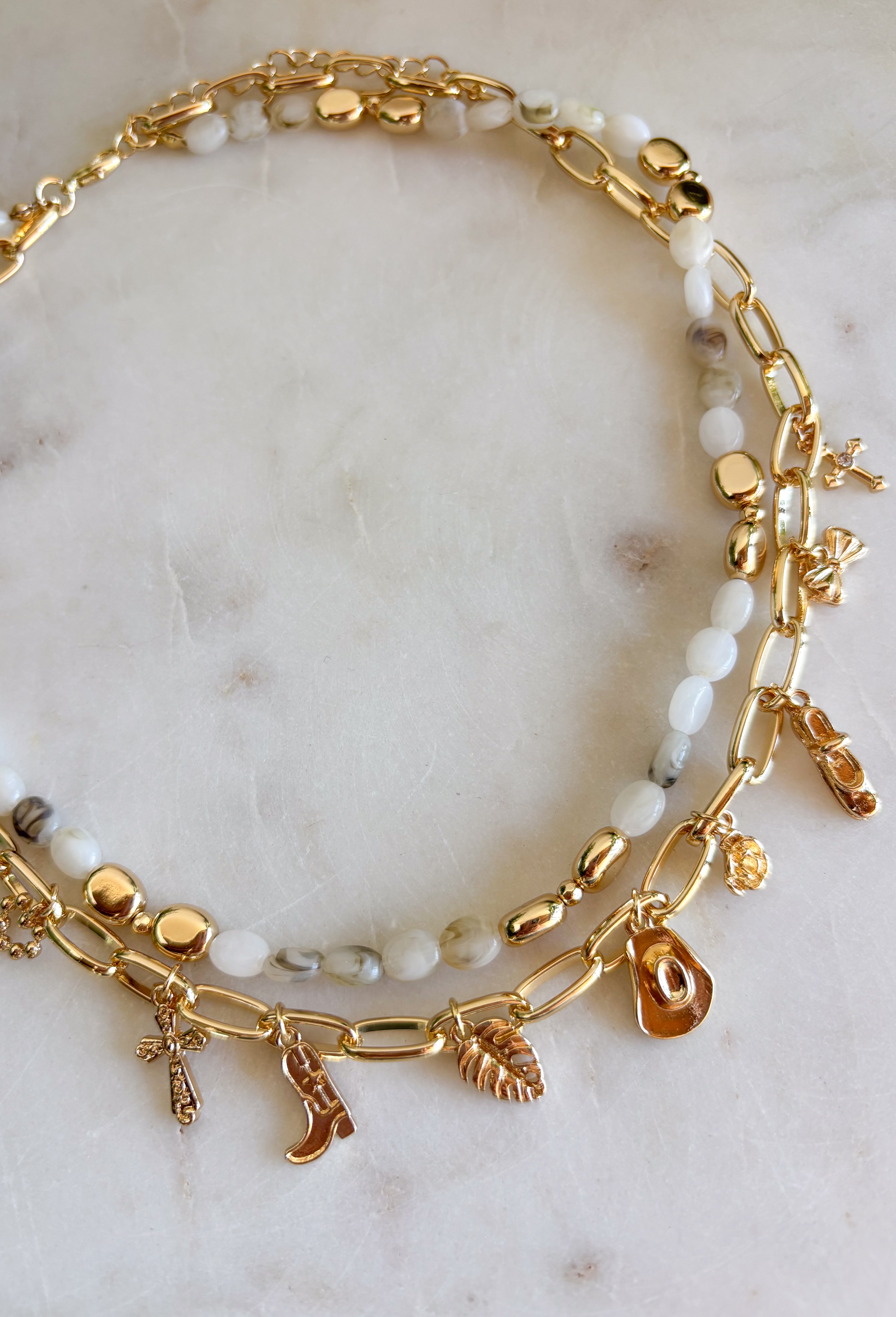 This layered necklace gives you that perfectly styled, on-trend look without any effort! Featuring a neutral beaded strand with gold accents paired with a charm-filled gold chain, it brings the perfect mix of western-inspired and playful details. With charms like a cross, heart, palm leaf, bow, and even a shoe, it adds so much personality to your outfit. Lightweight and easy to wear, it’s the ultimate throw-on accessory for summer days when you want to look put together in seconds. 