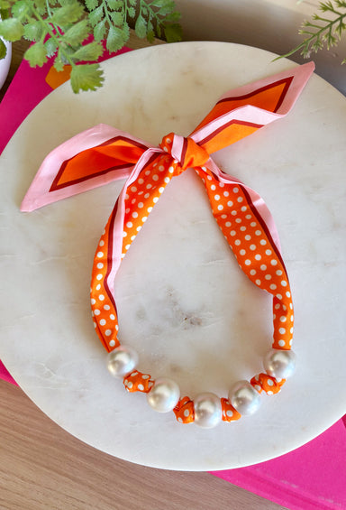 Add a playful pop of autumn color to your accessory collection with this versatile scarf necklace that's full of timeless charm. Featuring a cheerful orange polka dot print accented with brown and light pink trim, this unique piece is finished with chunky pearl detailing for a chic touch. The scarf tie design allows you to adjust the fit exactly how you like it, making it easy to wear as a necklace or wrap around your wrist as a chic bracelet.