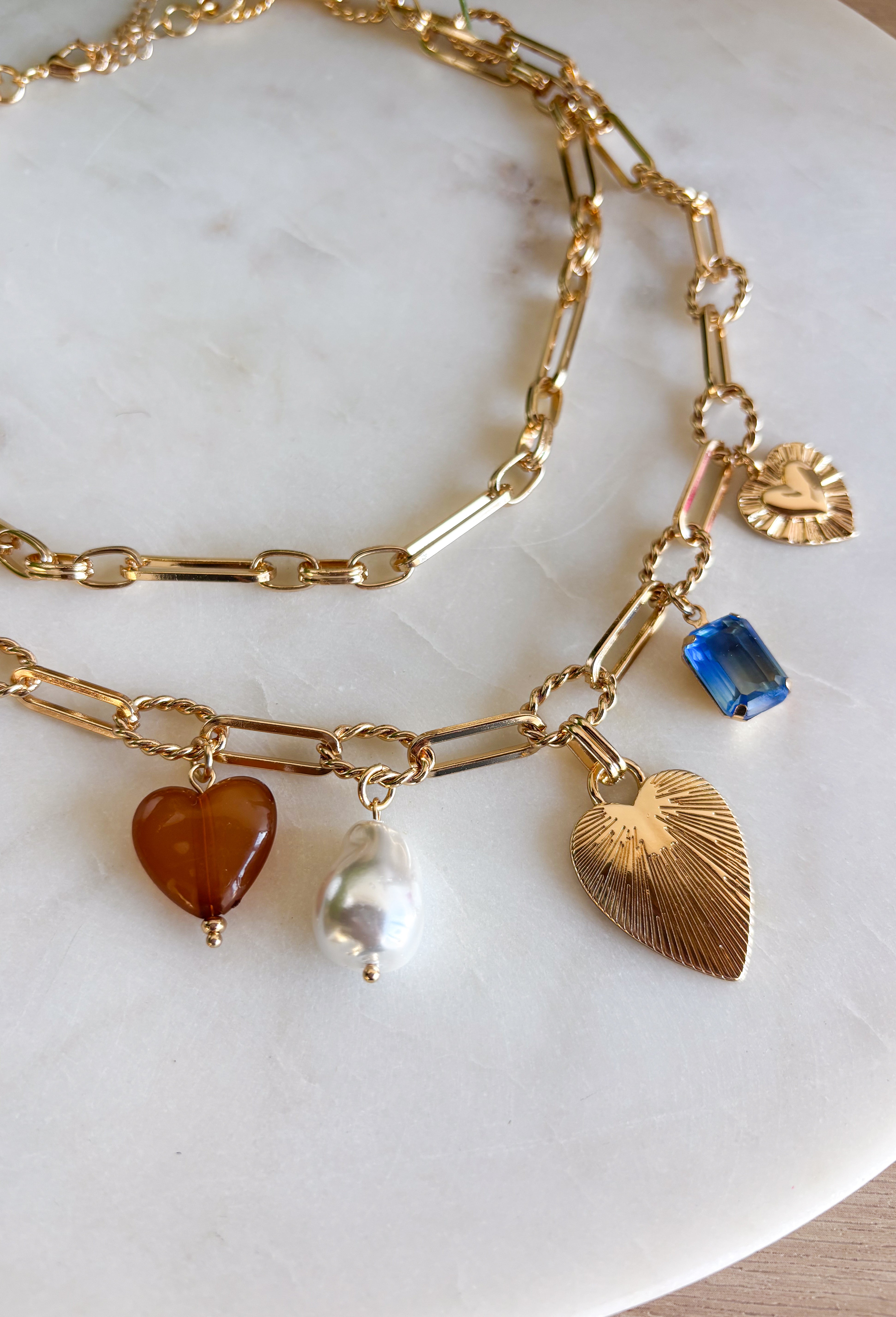 Turn any outfit into a statement with this chunky gold layered chain necklace. The double-layer design creates an effortless stacked look, while the second chain features an assortment of charming details — textured gold hearts, a pearl accent, a blue rhinestone, and a tan heart charm. It’s the perfect mix of bold and feminine, making it a go-to accessory for all season long.