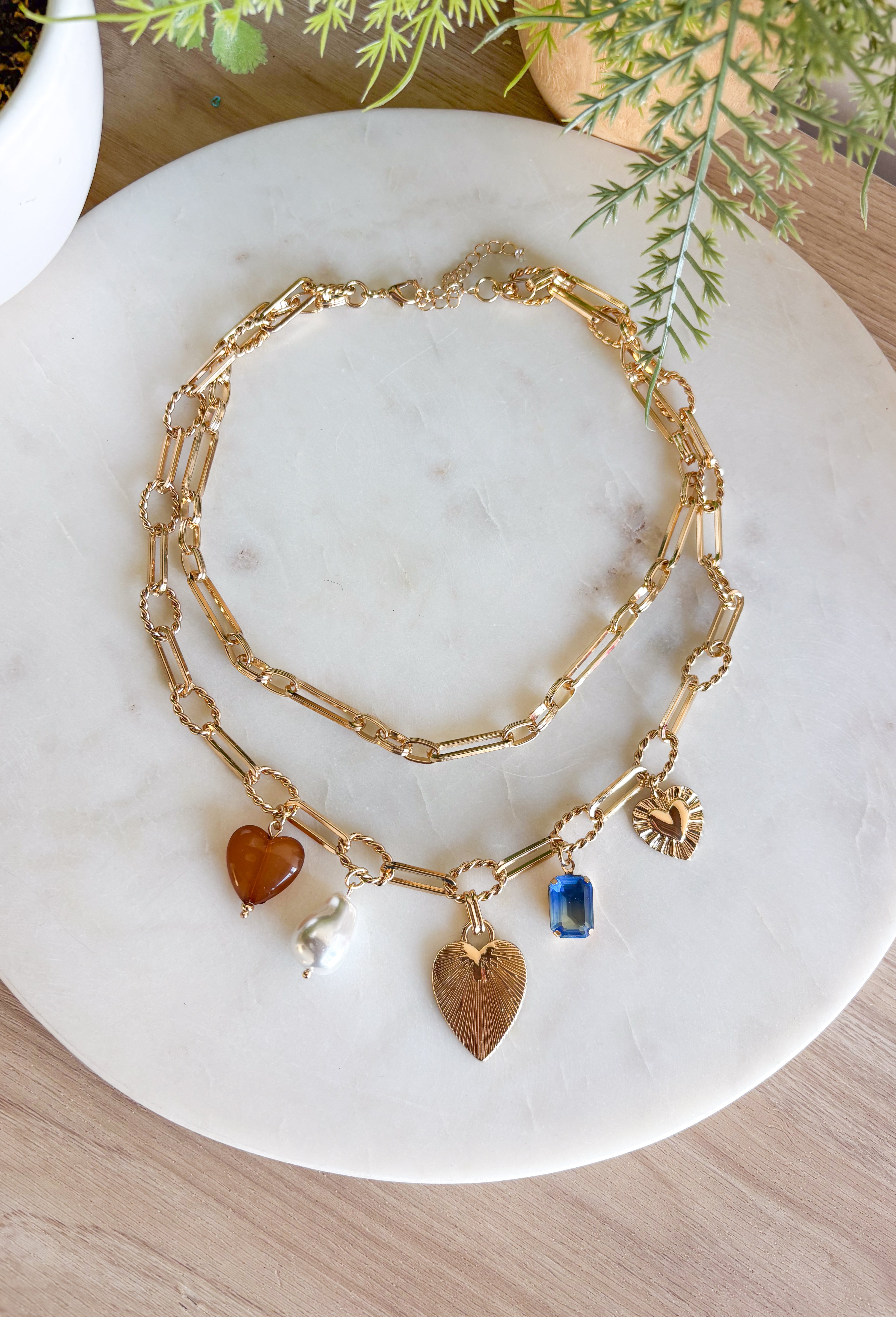 Turn any outfit into a statement with this chunky gold layered chain necklace. The double-layer design creates an effortless stacked look, while the second chain features an assortment of charming details — textured gold hearts, a pearl accent, a blue rhinestone, and a tan heart charm. It’s the perfect mix of bold and feminine, making it a go-to accessory for all season long.