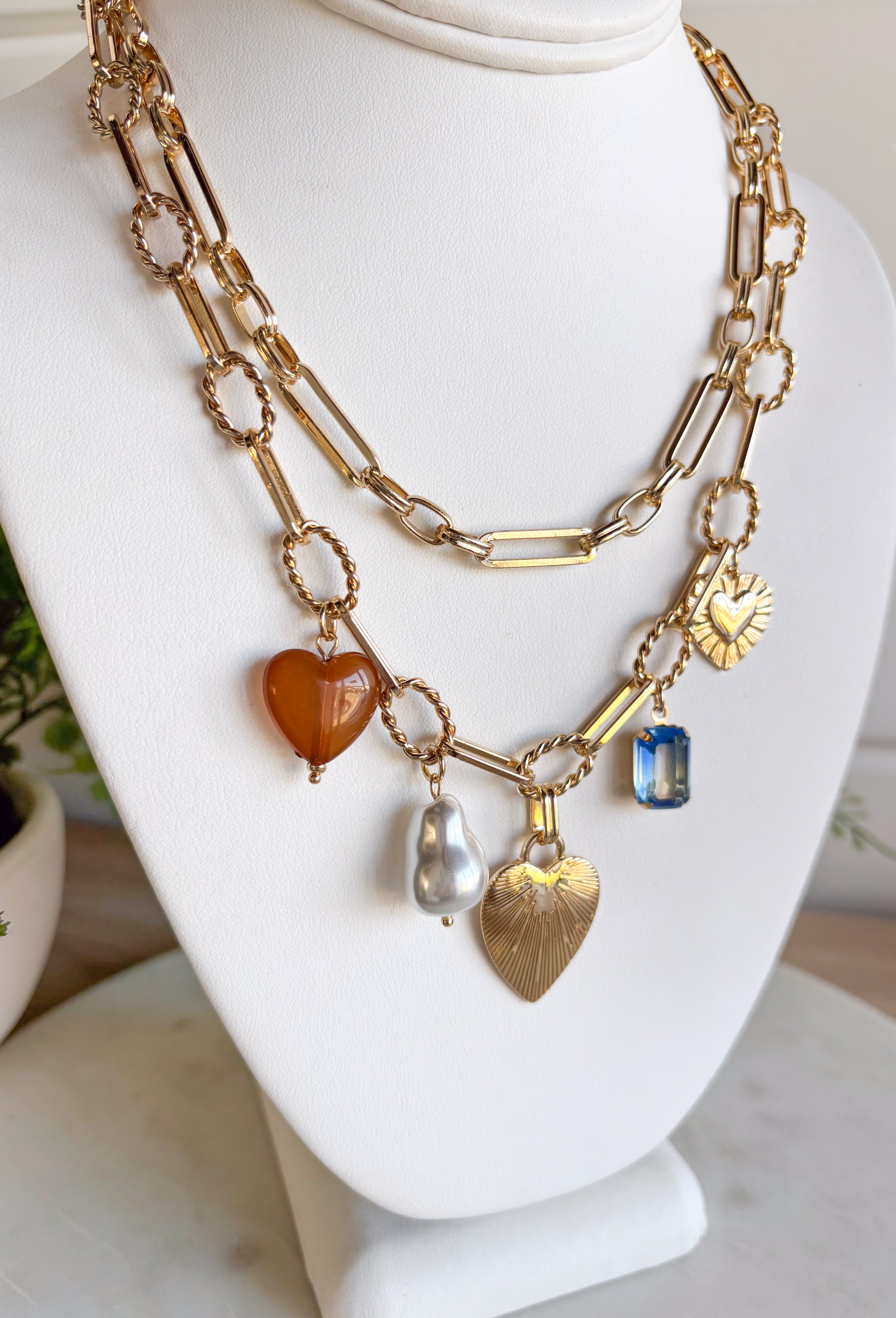 Turn any outfit into a statement with this chunky gold layered chain necklace. The double-layer design creates an effortless stacked look, while the second chain features an assortment of charming details — textured gold hearts, a pearl accent, a blue rhinestone, and a tan heart charm. It’s the perfect mix of bold and feminine, making it a go-to accessory for all season long.