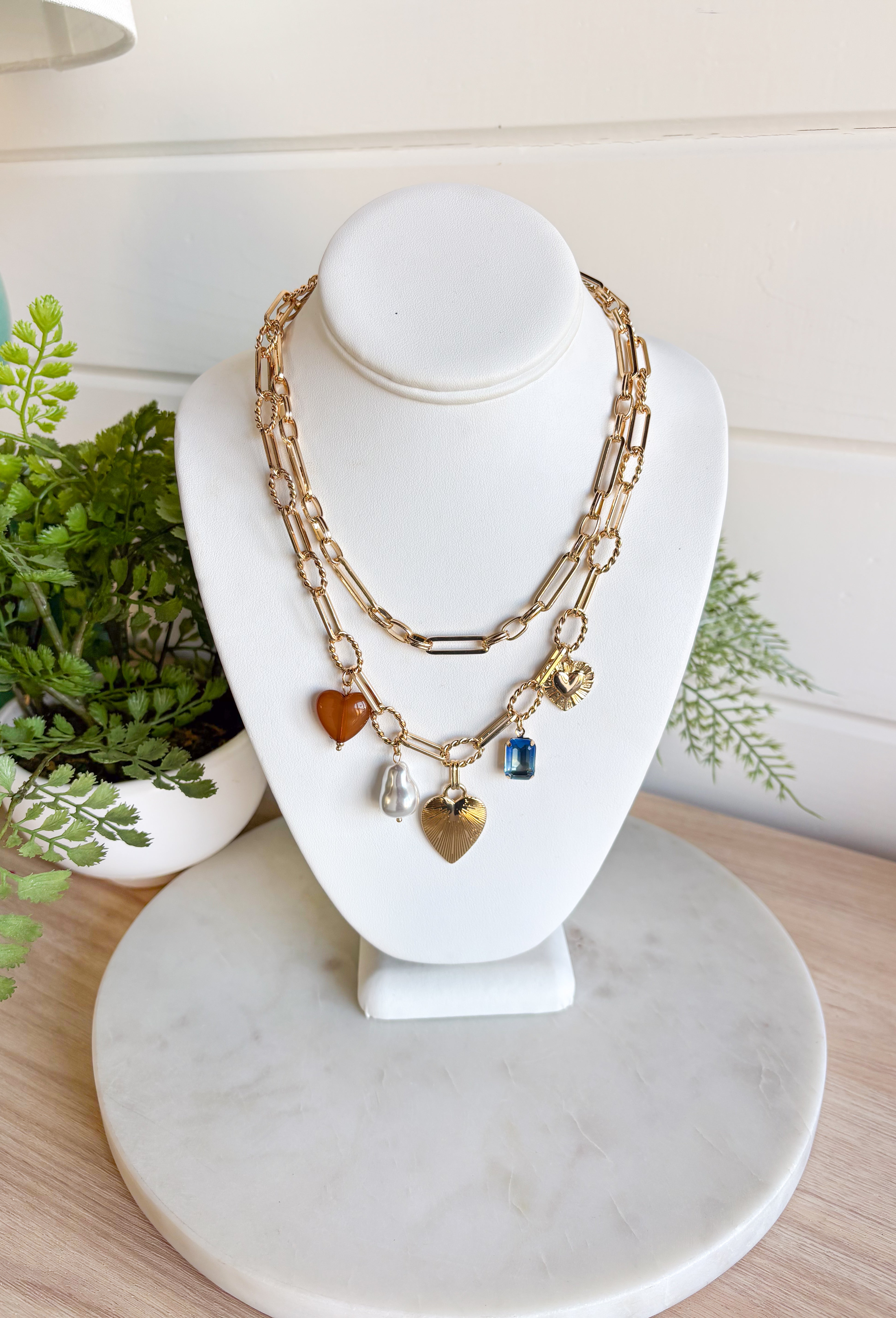 Turn any outfit into a statement with this chunky gold layered chain necklace. The double-layer design creates an effortless stacked look, while the second chain features an assortment of charming details — textured gold hearts, a pearl accent, a blue rhinestone, and a tan heart charm. It’s the perfect mix of bold and feminine, making it a go-to accessory for all season long.